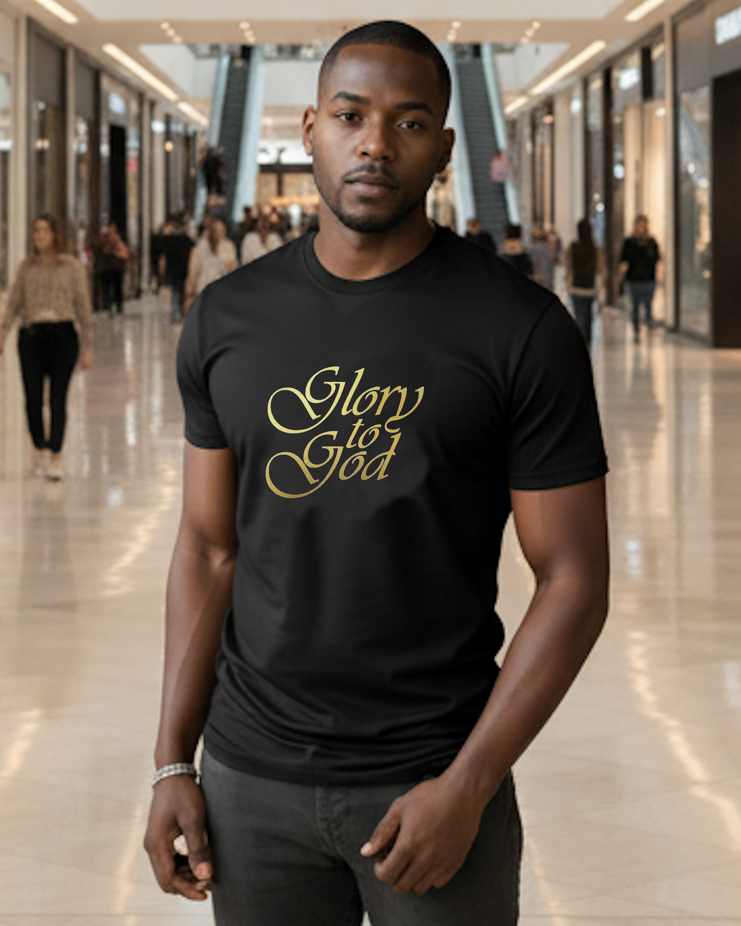 A man wearing a black crewneck t-shirt with 'Glory to God' printed in gold cursive script, standing in a brightly lit, blurred shopping mall corridor.
