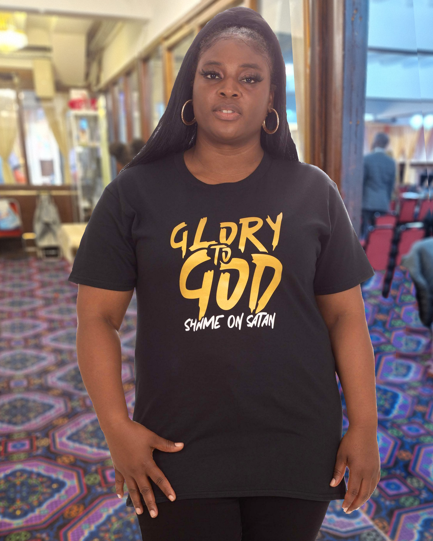 A woman in a church wearing a black t-shirt with a gold Heavenly Dreamwear "Glory to God" logo printed on the front.