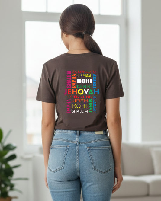 A woman wearing a dark brown t-shirt with a colorful rainbow text graphic on the back that reads 'JEHOVAH, TSIDKENU, SHAMMAH, SHALOM, JIREH, RAPHA NISSI, ROHI' paired with light blue jeans, standing in a bright, modern room.