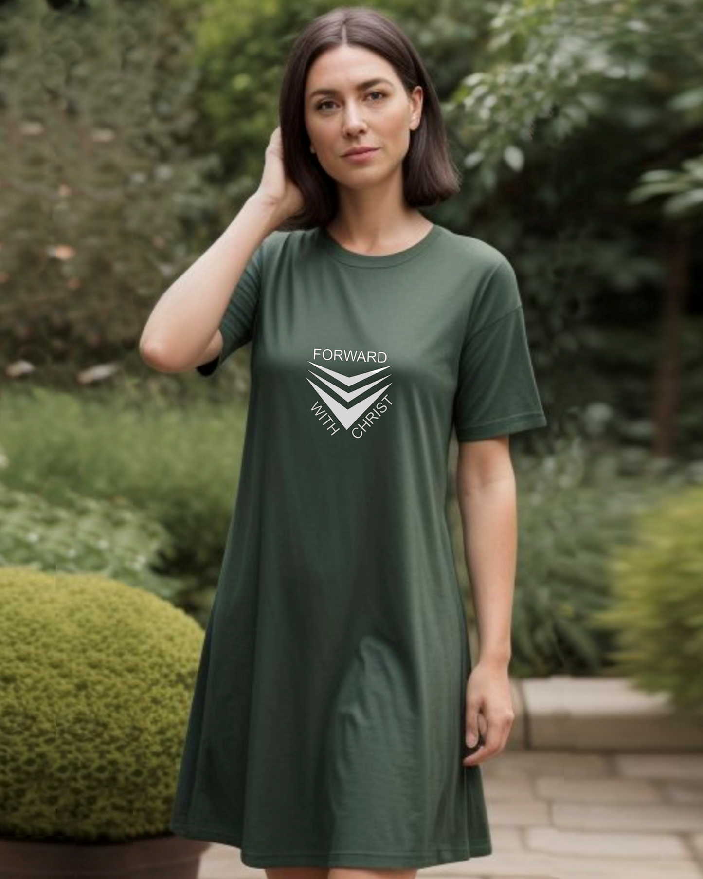 A woman standing in a garden wearing a dark green Heavenly Dreamwear t-shirt dress with a white logo made up of 3 chevrons surrounded by the text, "Forward With Christ".