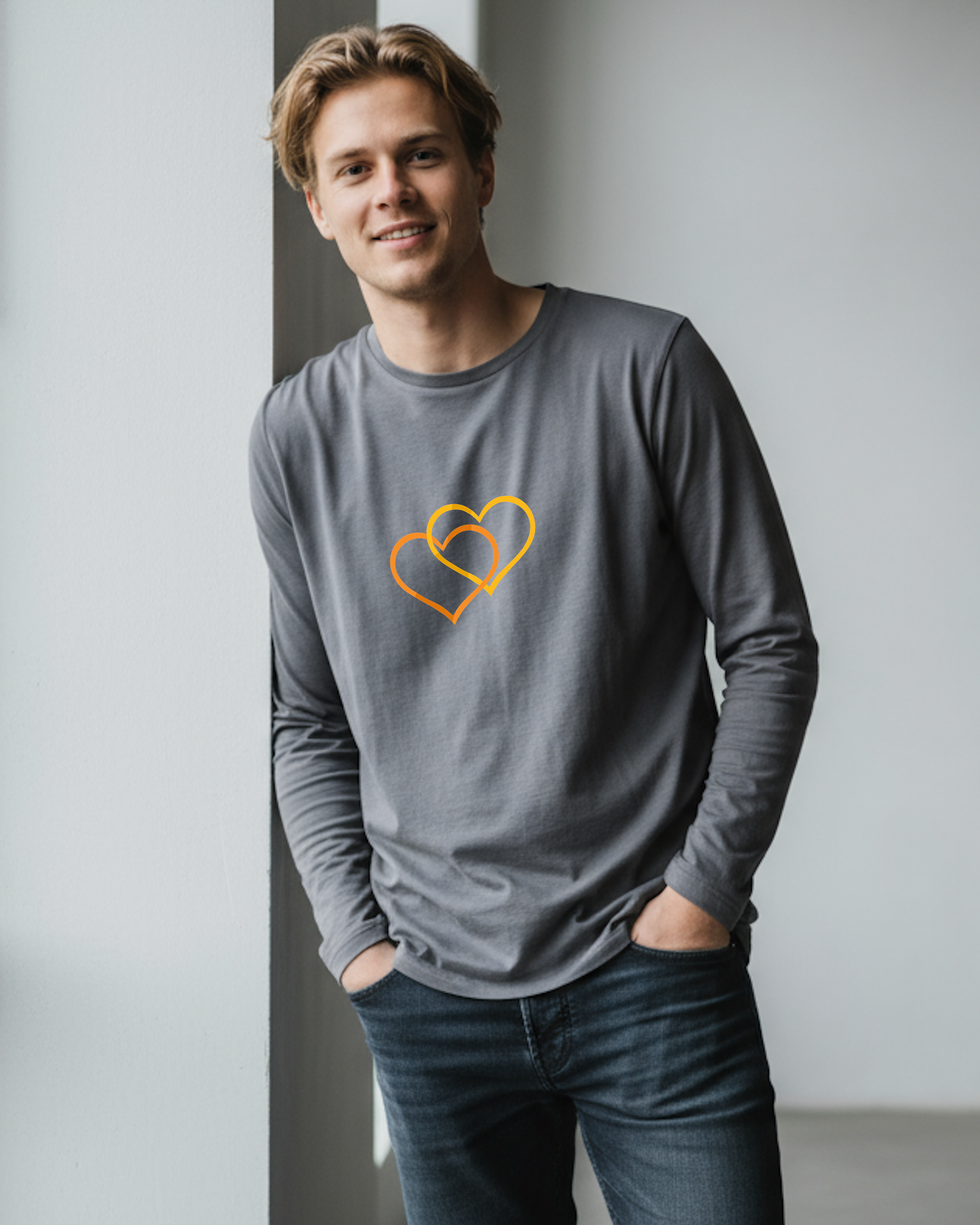 Man wearing a gray long-sleeve shirt with an orange heart interleaved with a yellow heart design leaning against grey wall.