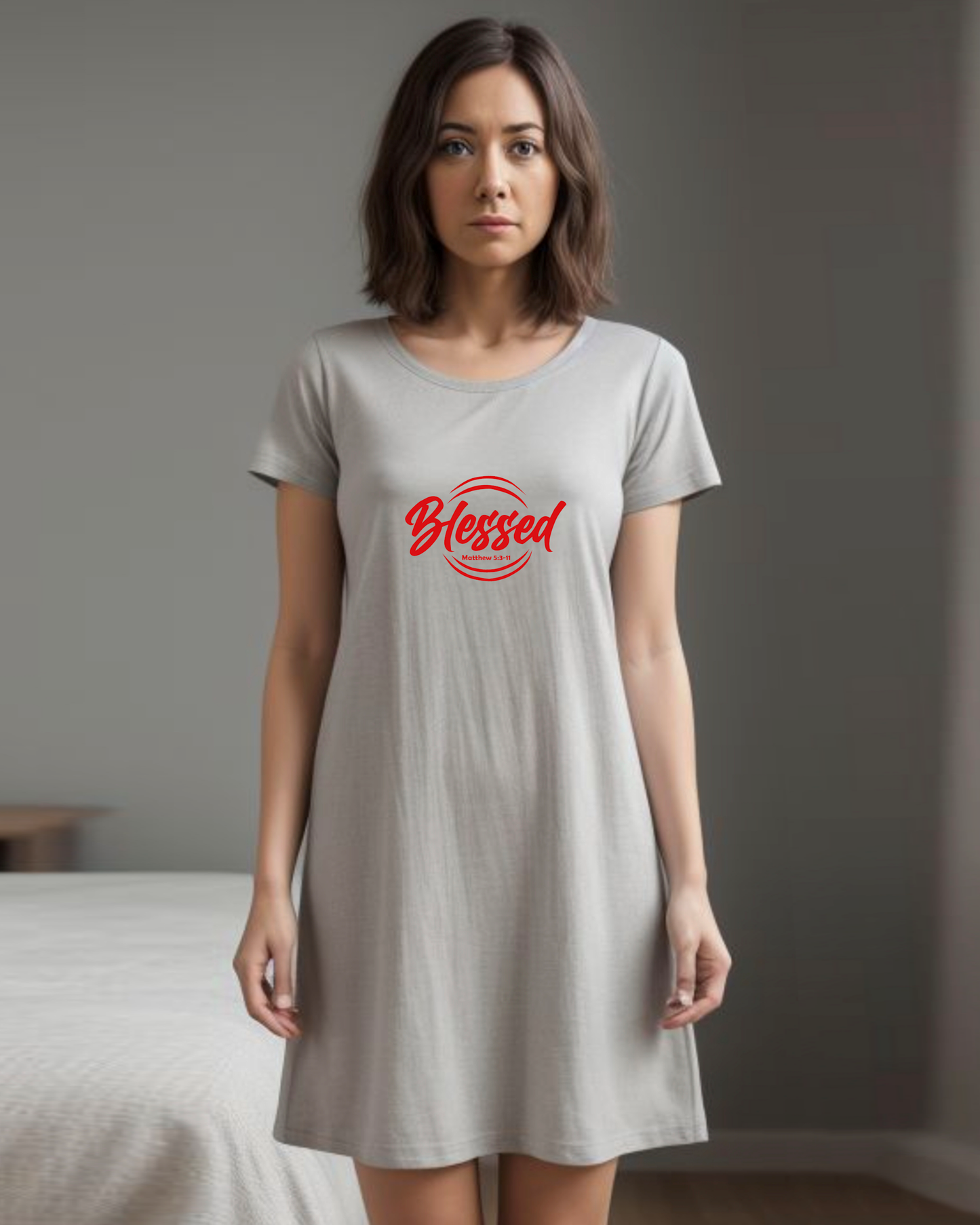 A woman in a bedroom wearing a grey Heavenly Dreamwear t-shirt dress with a red logo comprised of the text "Blessed" on top of a broken double circle with a biblical scripture reference beneath the text.
