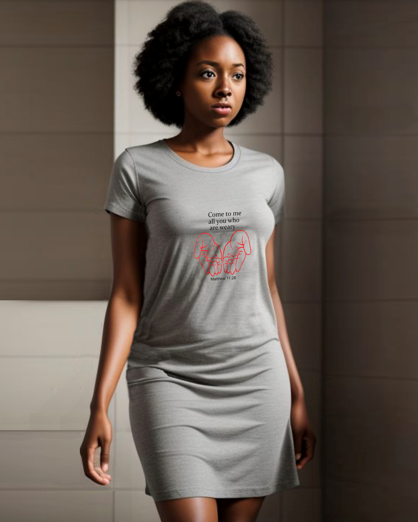 A woman in a bathroom wearing a grey Heavenly Dreamwear t-shirt dress with a black "come to me all you who are weary" text with two red hands facing palm up logo printed on the front.