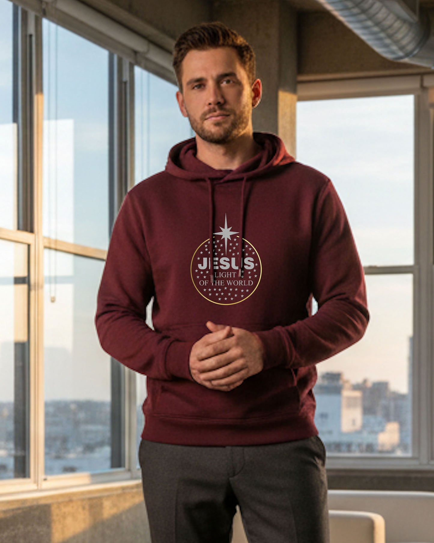 Man wearing a maroon hoodie with a logo standing in front of large windows with a cityscape view.