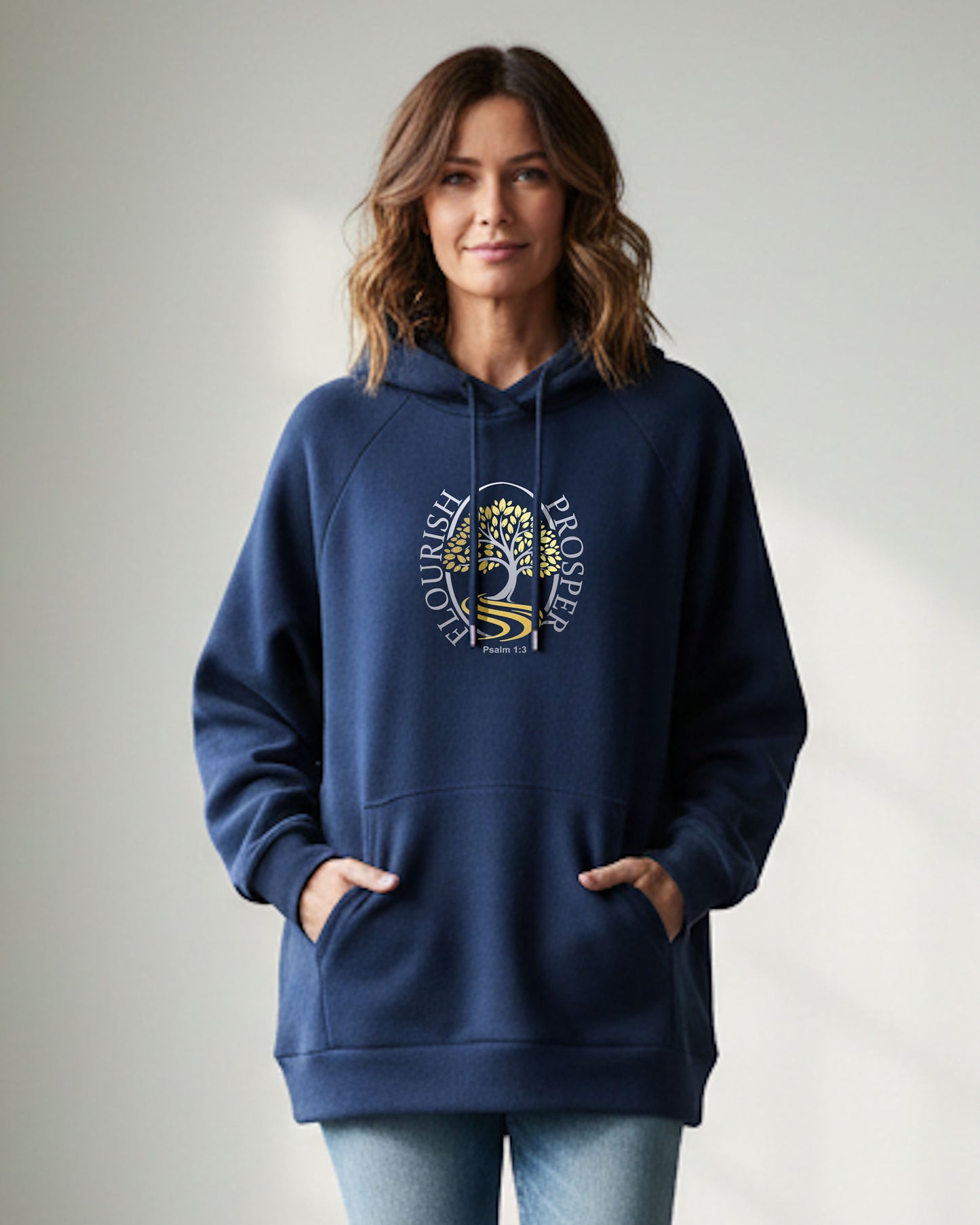 Woman leaning on a wall wearing a navy blue hoodie with a gold/silver logo with text 'Flourish Prosper Psalm 1:3' against a plain backgound