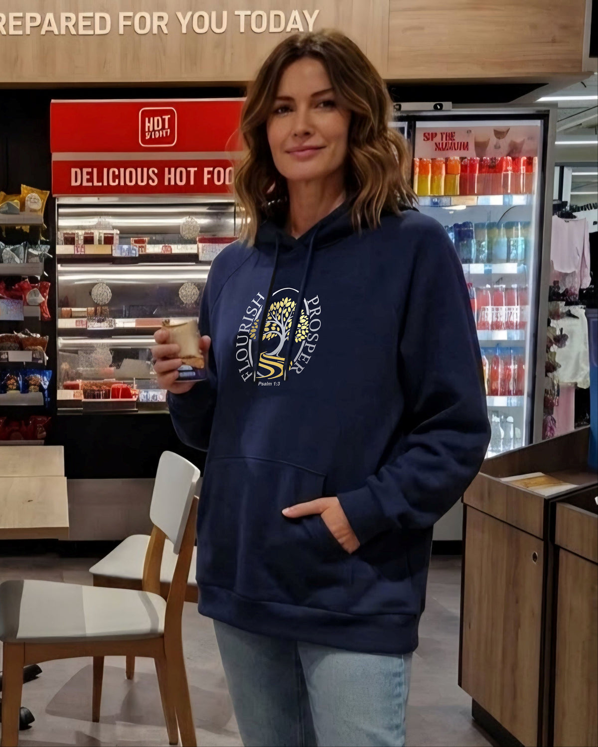 A woman wearing a navy blue hoodie with a 'Flourish Prosper' silver and gold graphic logo stands inside a retail food environment, holding a cup.