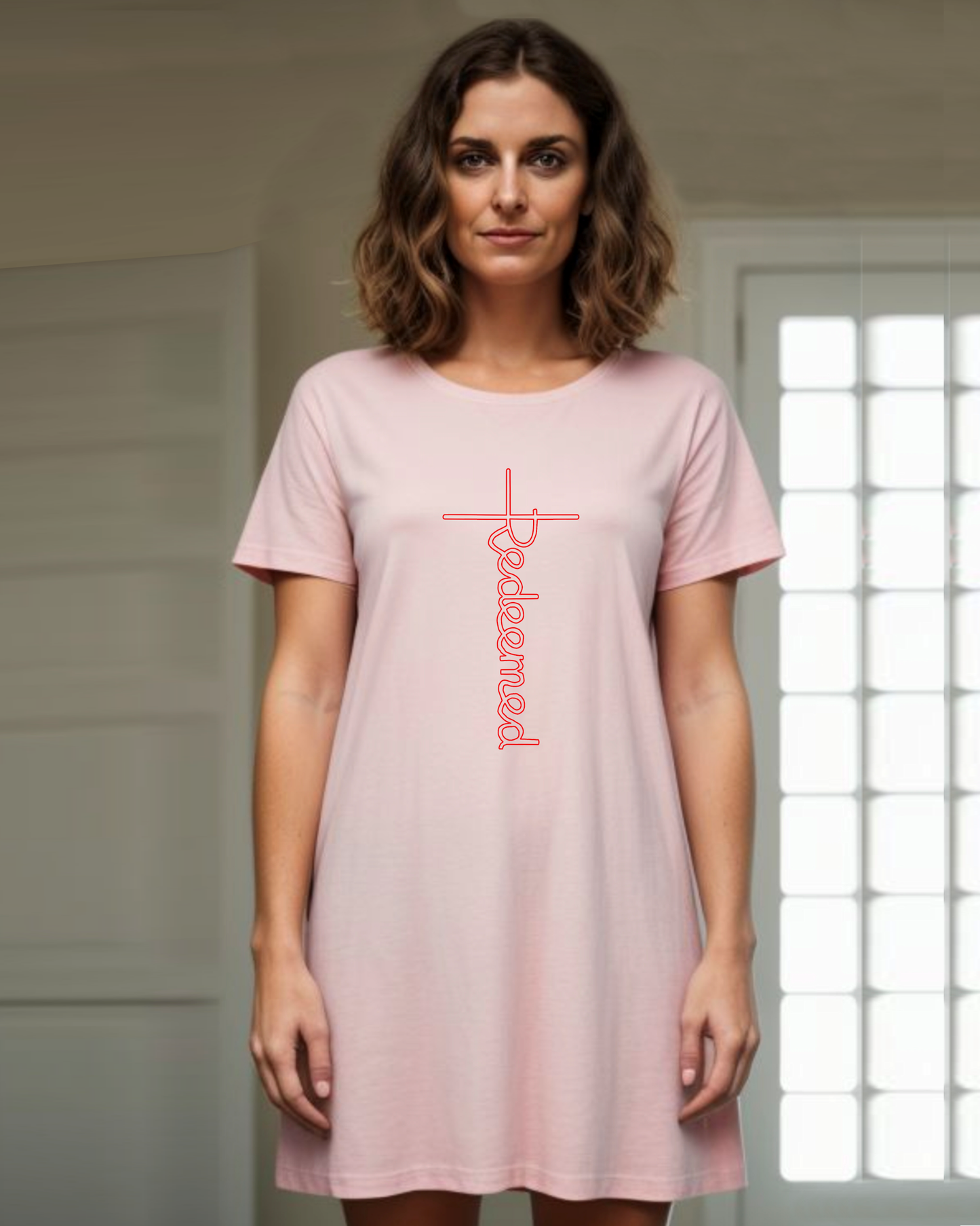 A woman in a bedroom wearing a pink T-shirt dress with an elaborate red Heavenly Dreamwear designed 'Redeemed' logo printed on the front.