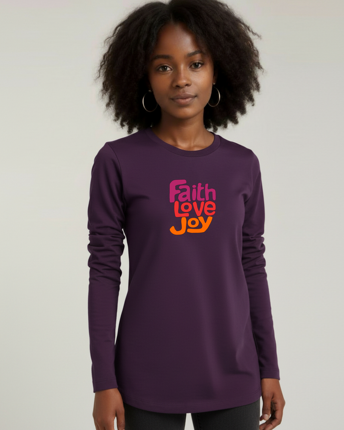 A woman with curly black hair wearing a purple long-sleeve t-shirt with a colorful 'Faith Love Joy' graphic on the chest, set against a plain light gray background.