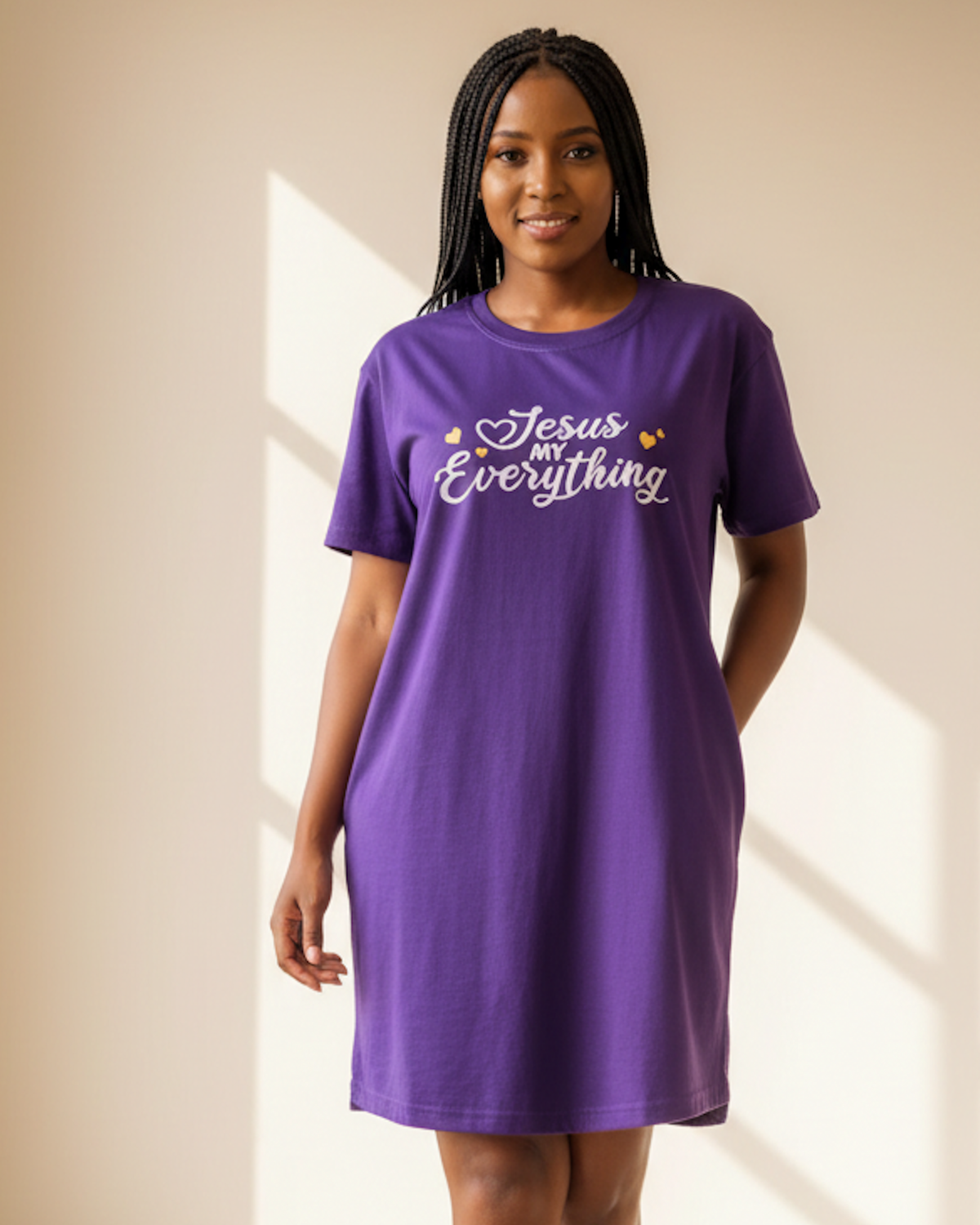 A woman modeling a purple short-sleeve t-shirt dress with the text 'Jesus My Everything' in white script and small yellow hearts on the chest, set against a warm beige background with soft window shadows.