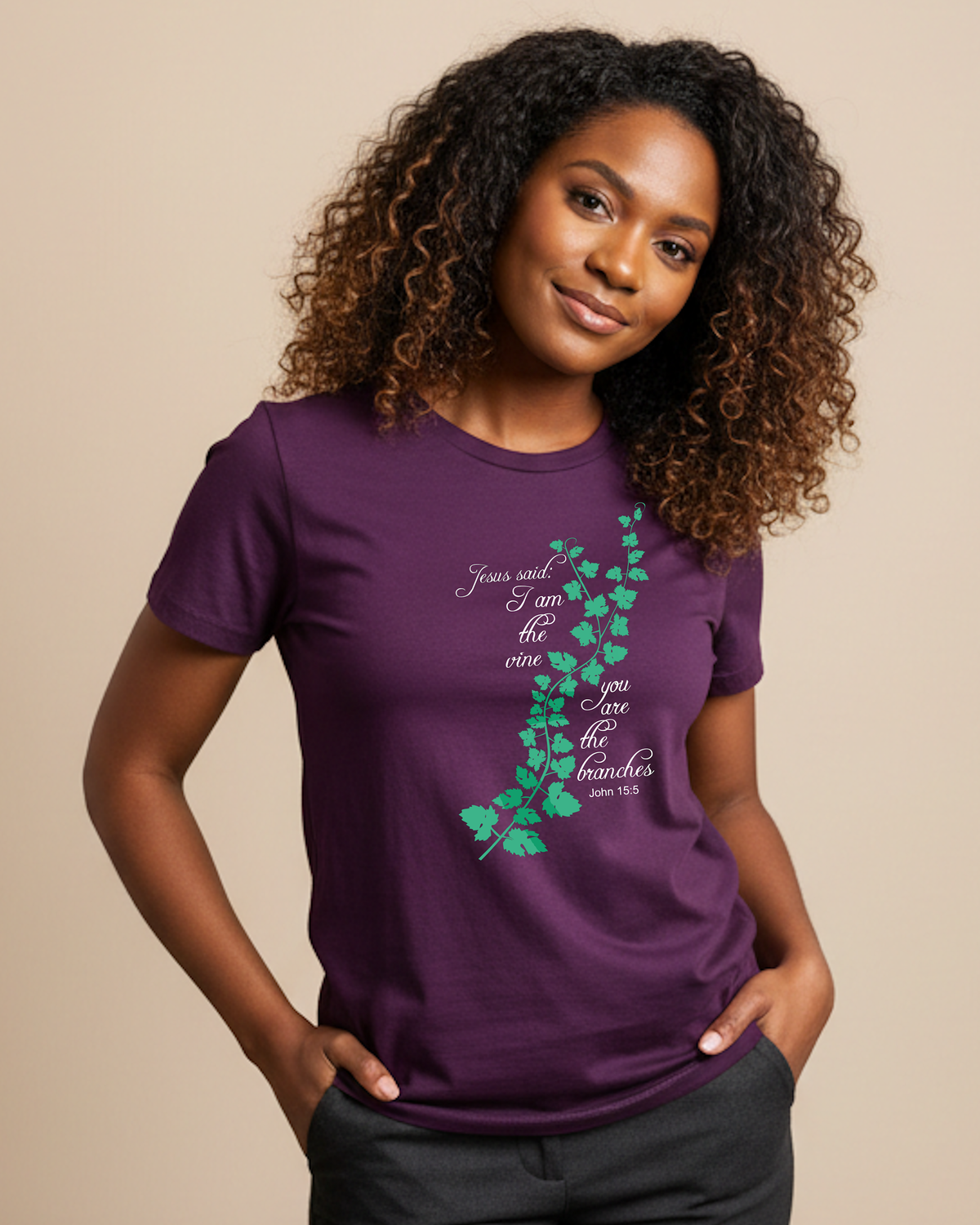 A woman with curly brown hair wearing a purple short-sleeve t-shirt with a green ivy vine graphic and white scripture text 'Jesus said I am the vine you are the branches' on the front, paired with dark gray pants, against a solid beige background.