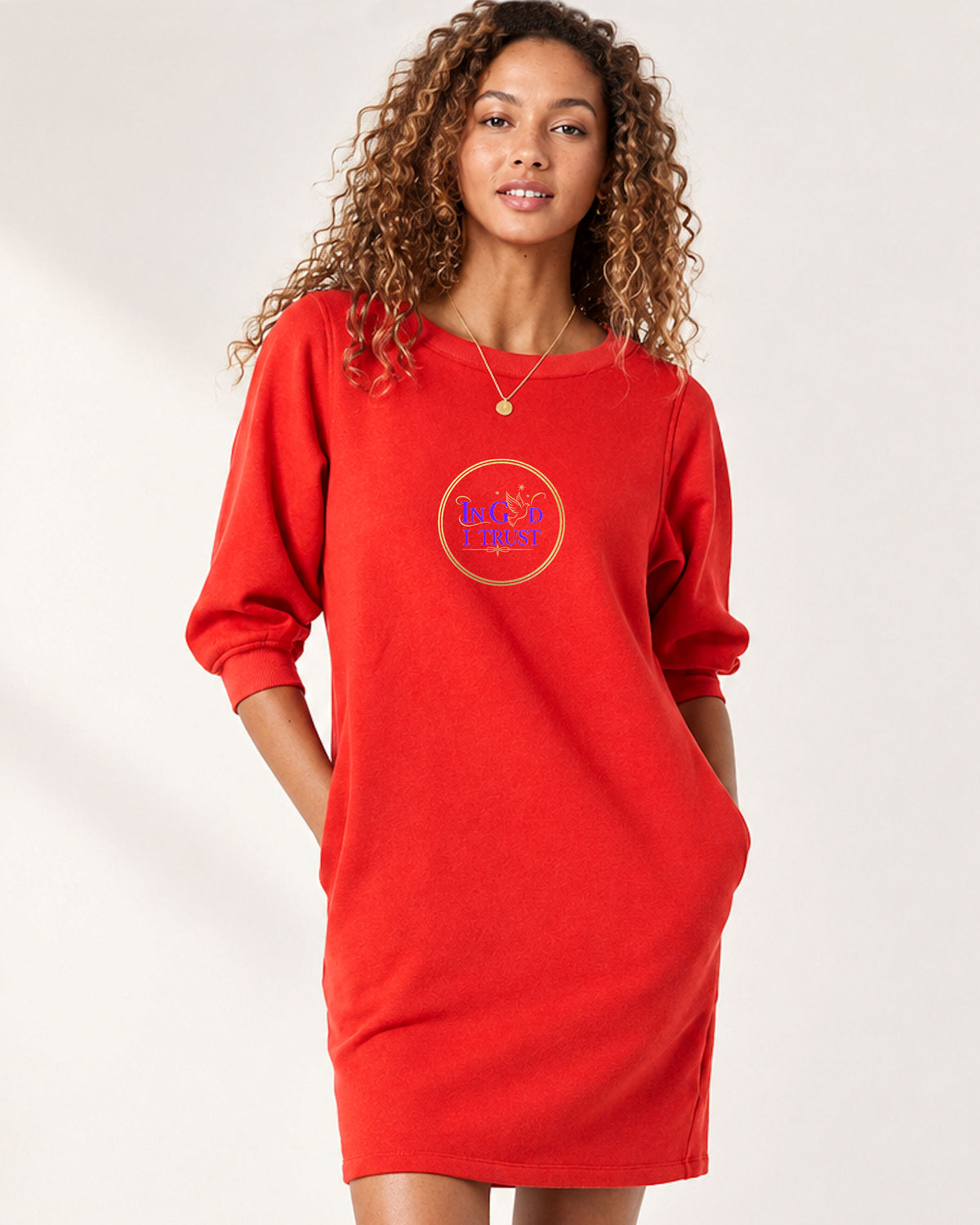 A woman modeling a bright red sweatshirt dress with three-quarter sleeves and a circular chest graphic that reads 'In God I Trust' in purple and gold, set against a plain light background.