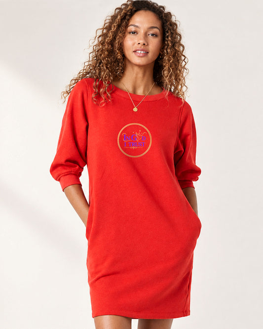 A woman modeling a bright red sweatshirt dress with three-quarter sleeves and a circular chest graphic that reads 'In God I Trust' in purple and gold, set against a plain light background.