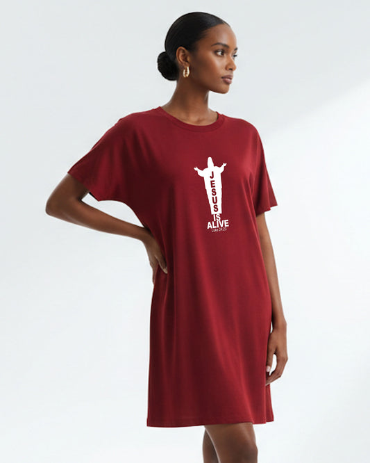 A woman wearing a red Heavenly Dreamwear t-shirt dress with a white logo of a silhouette of Jesus with the name "JESUS" embedded in it. Underneath the logo is the text "IS ALIVE" with the scriptural reference: Luke 24:23. Photo taken against a white wall.