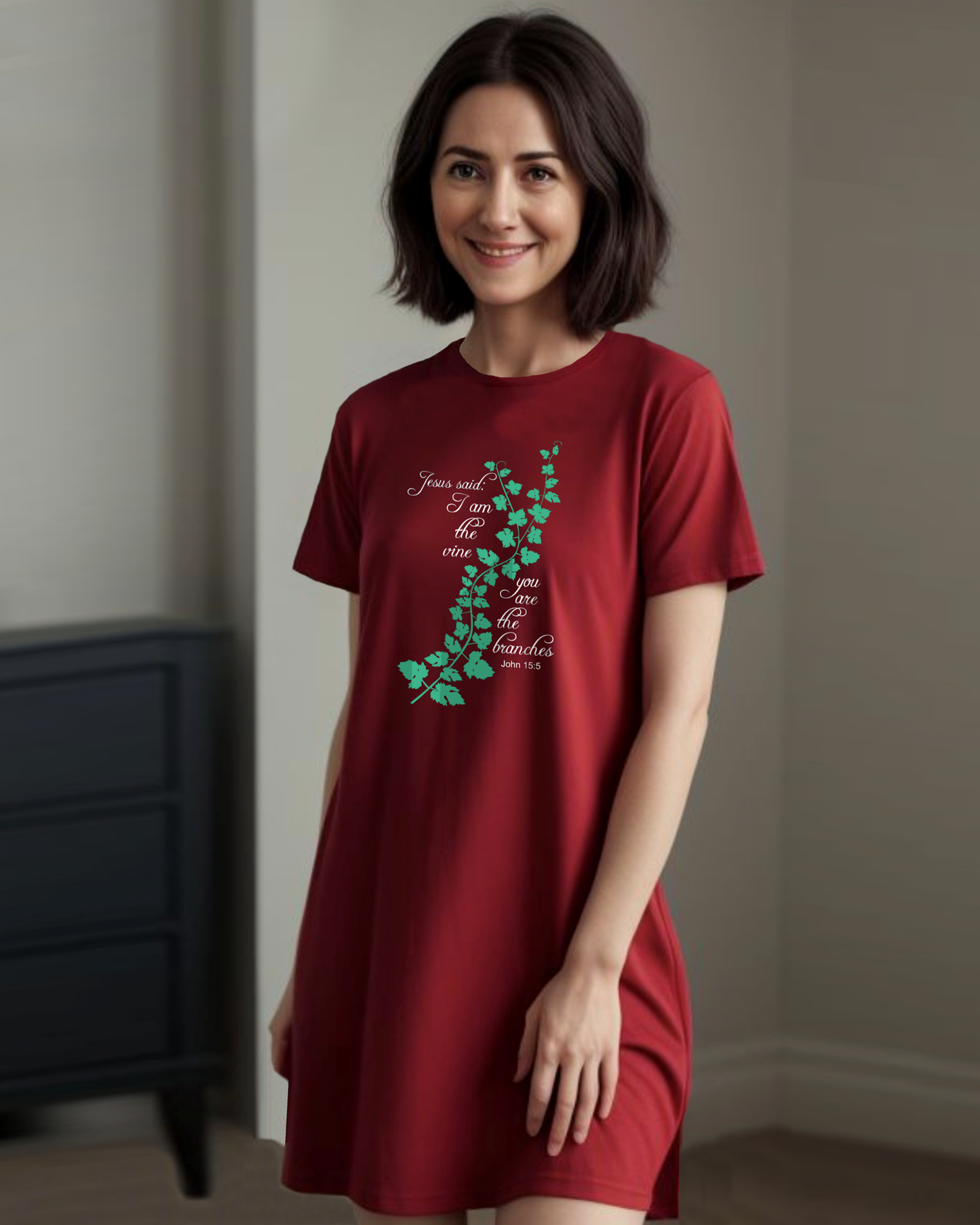 Woman standing in a bedroom wearing a red t-shirt dress with a logo made up of a green vine draped across the front with a biblical scripture on either side in white text from John 15:5