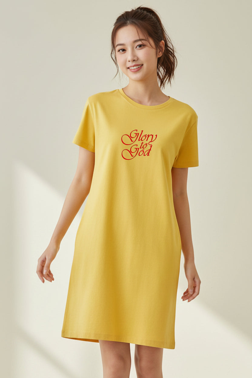 A woman indoors wearing a yellow t-shirt dress with red text the says, 'Glory to God' against a pale yellow wall