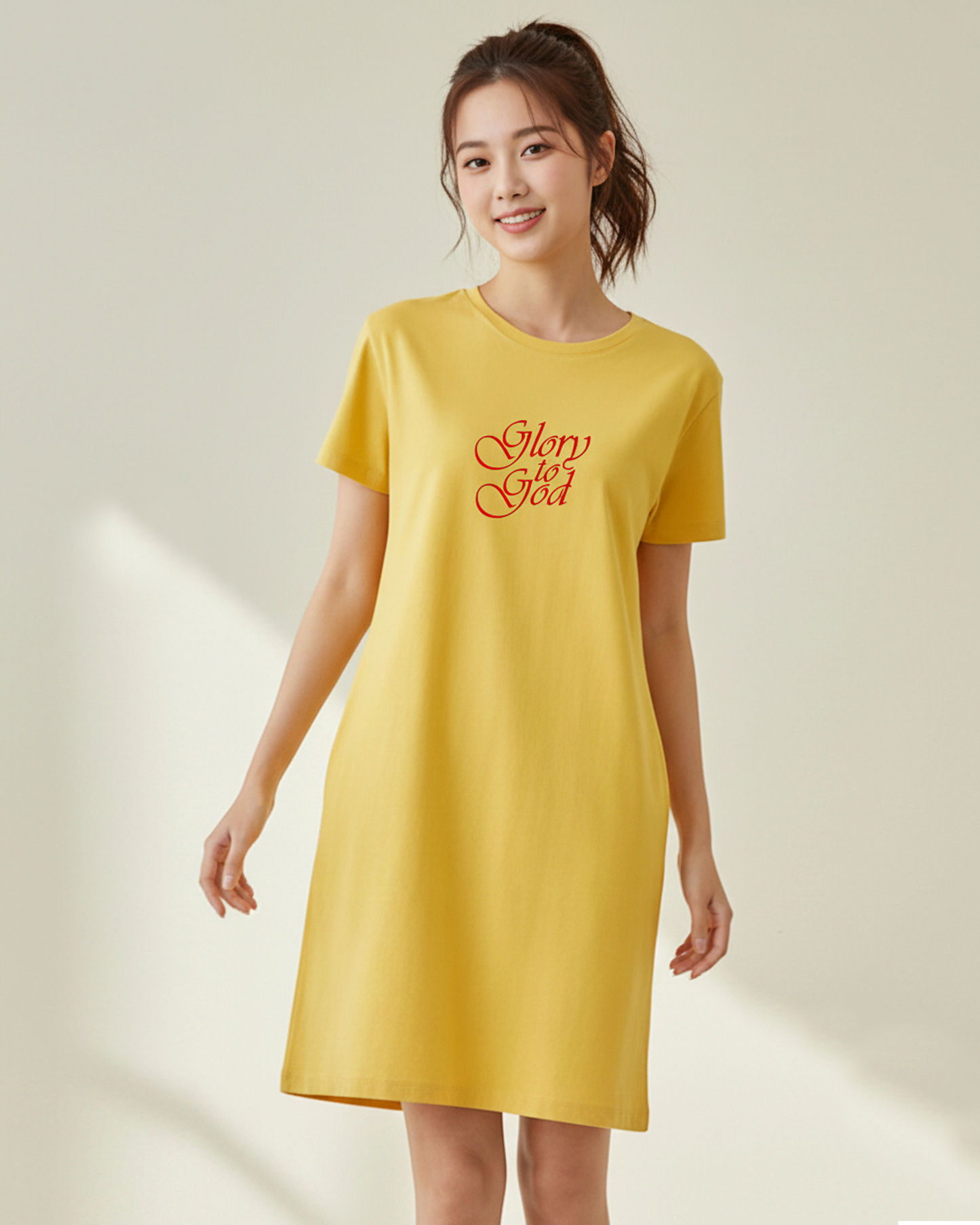 A woman wearing a yellow t-shirt dress with a Heavenly Dreamwear logo made of an purple and blue intertwined heart shapes on the front against a plain beige background.