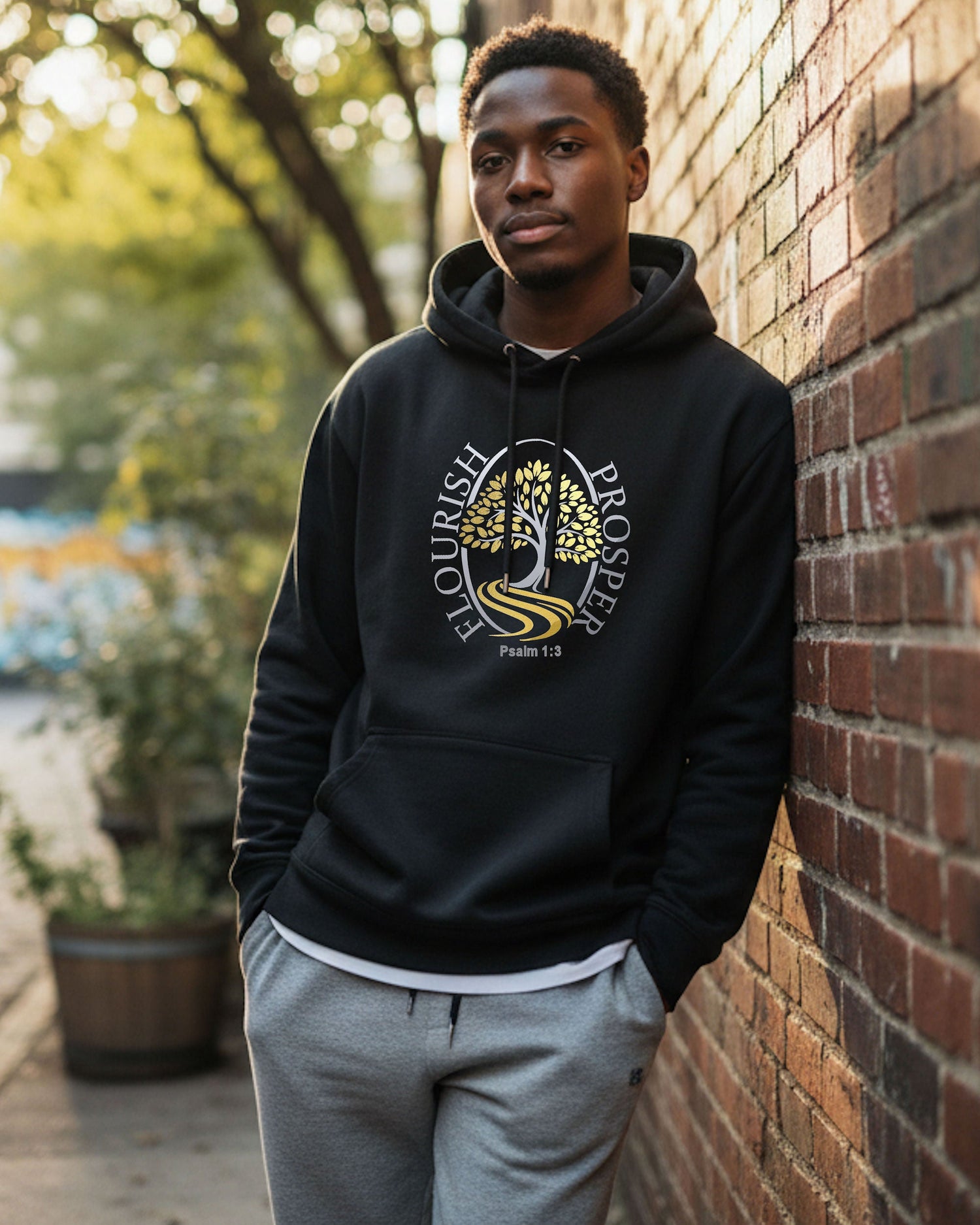 A young man leaning against a wall wearing a black hoodie with a gold/silver logo and text, 'Flourish Prosper'