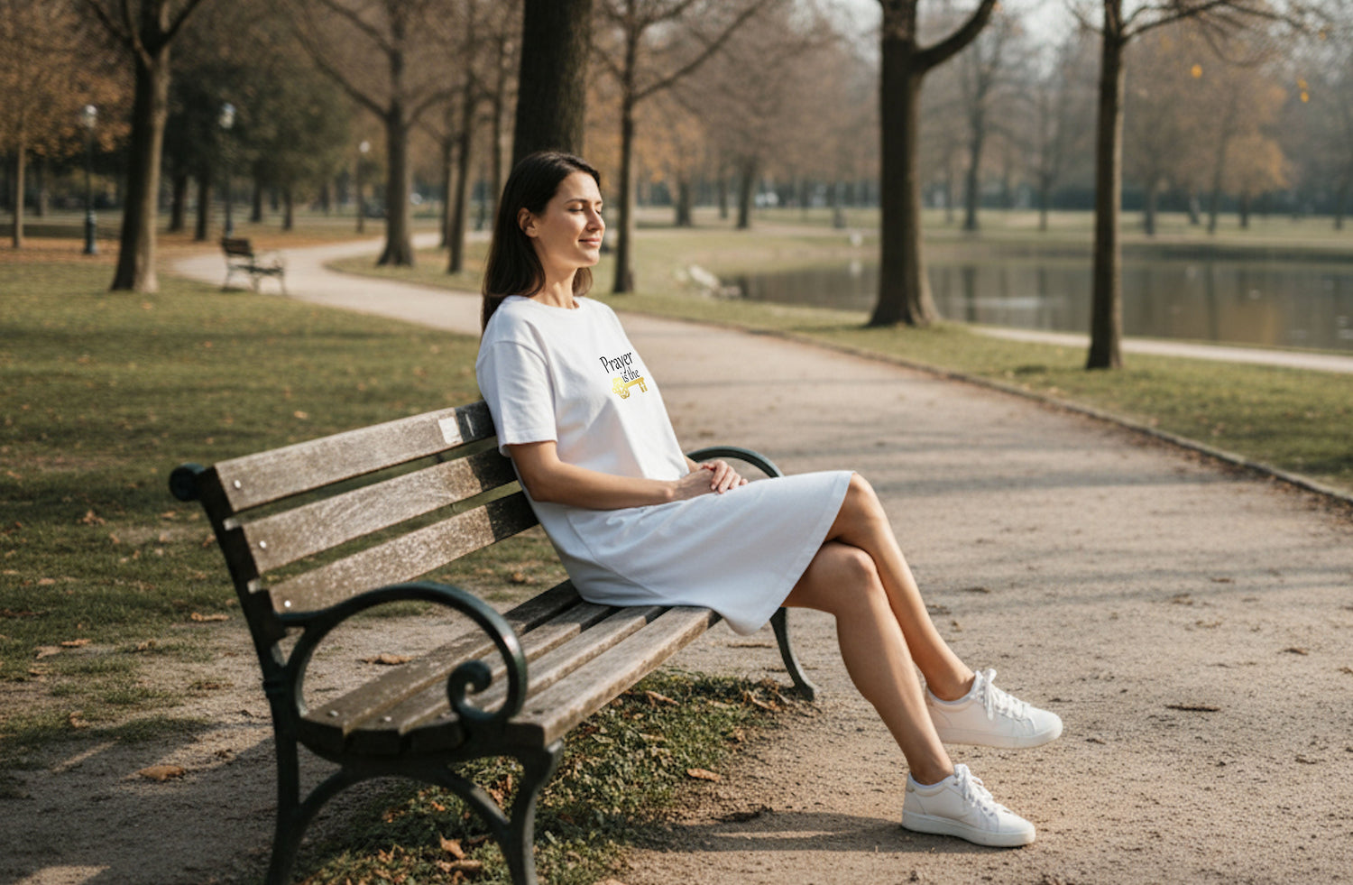 A woman sitting on a bench in a park wearing a white t-shirt dress
