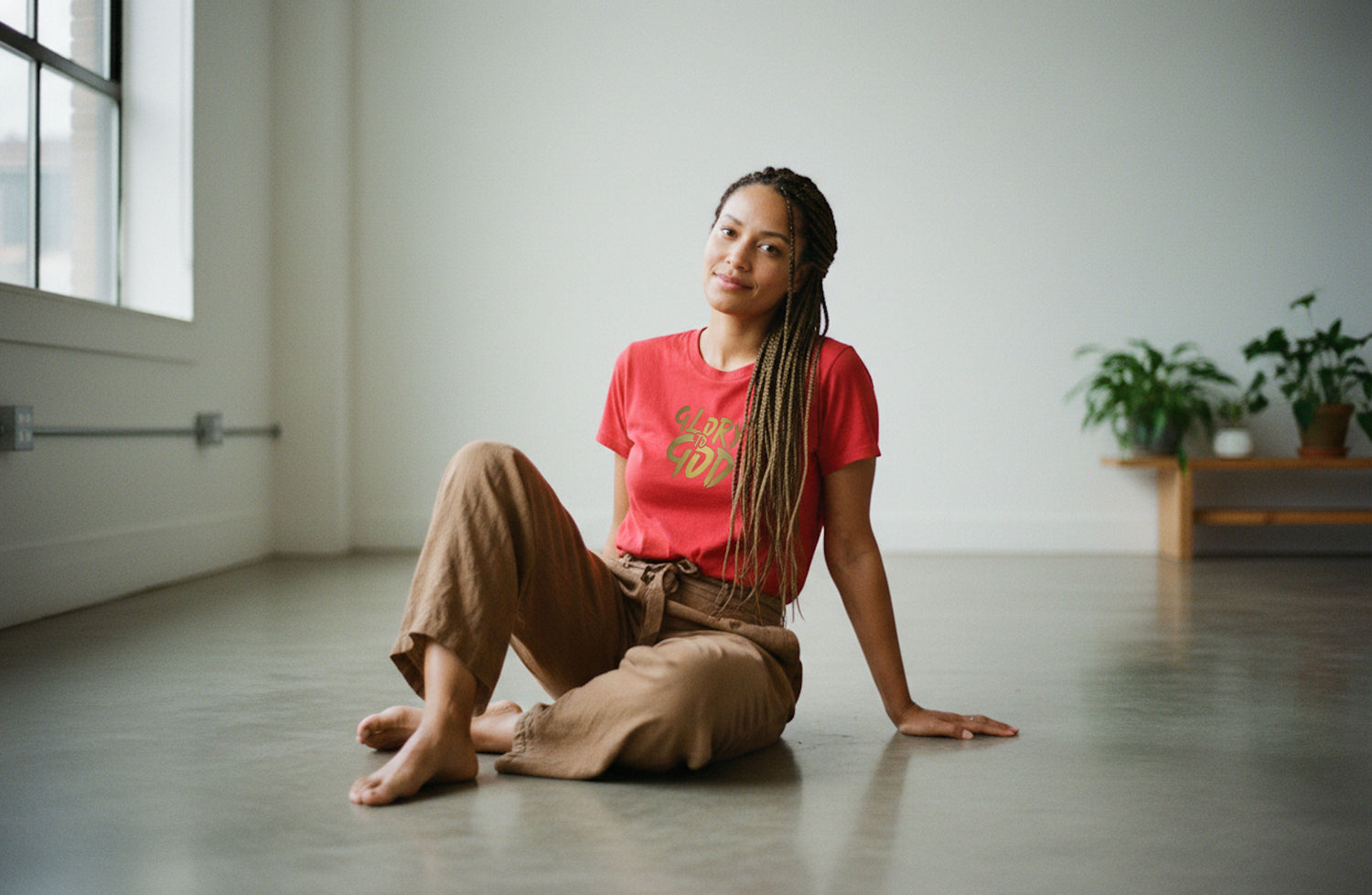 A woman sitting alone on a floor in an empty room, wearing a red short  sleeve t-shirt with a gold "Glory to God" logo printed on the front.