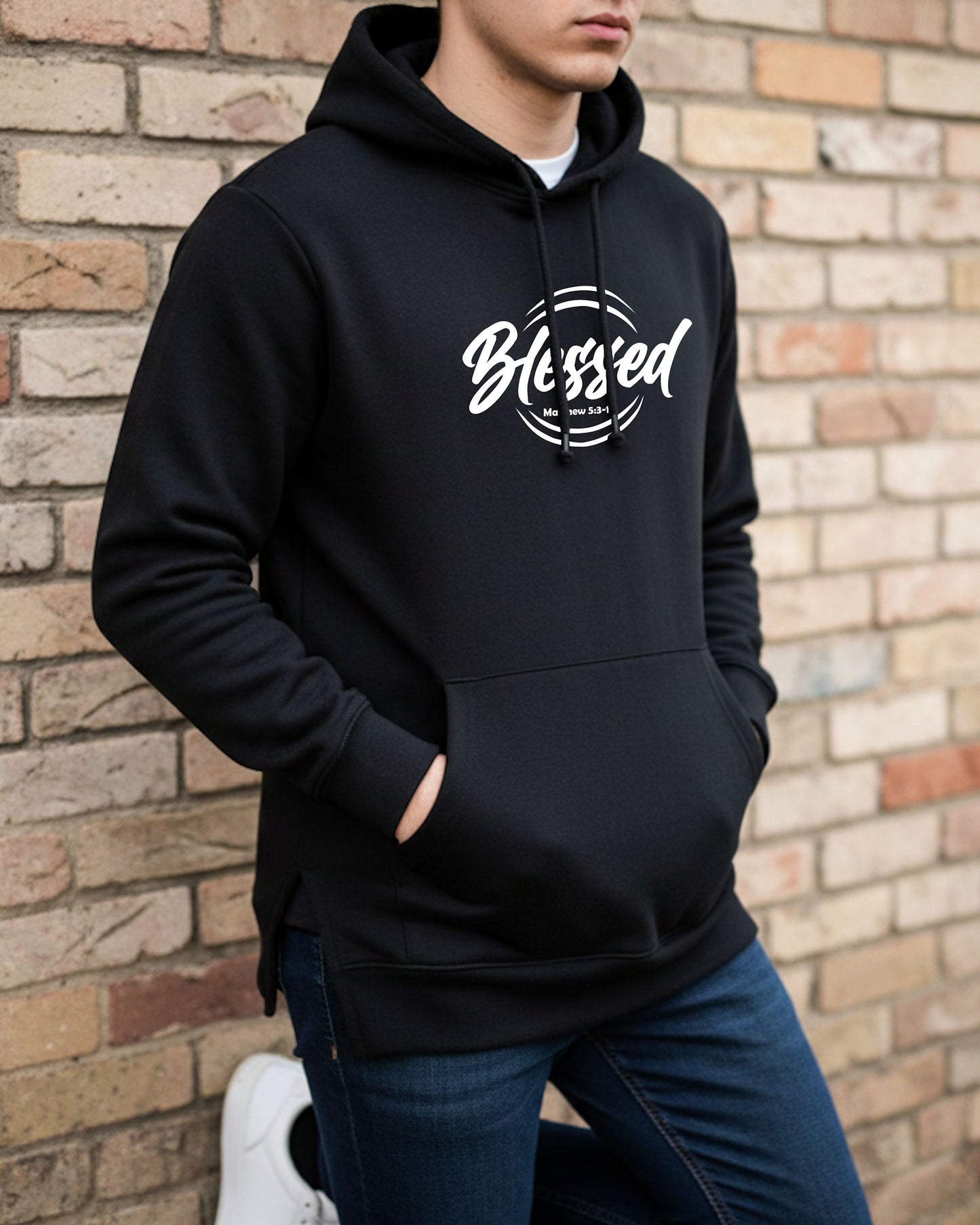 A young man leaning against a brick wall wearing a black hoodie with a text logo that says "Blessed"