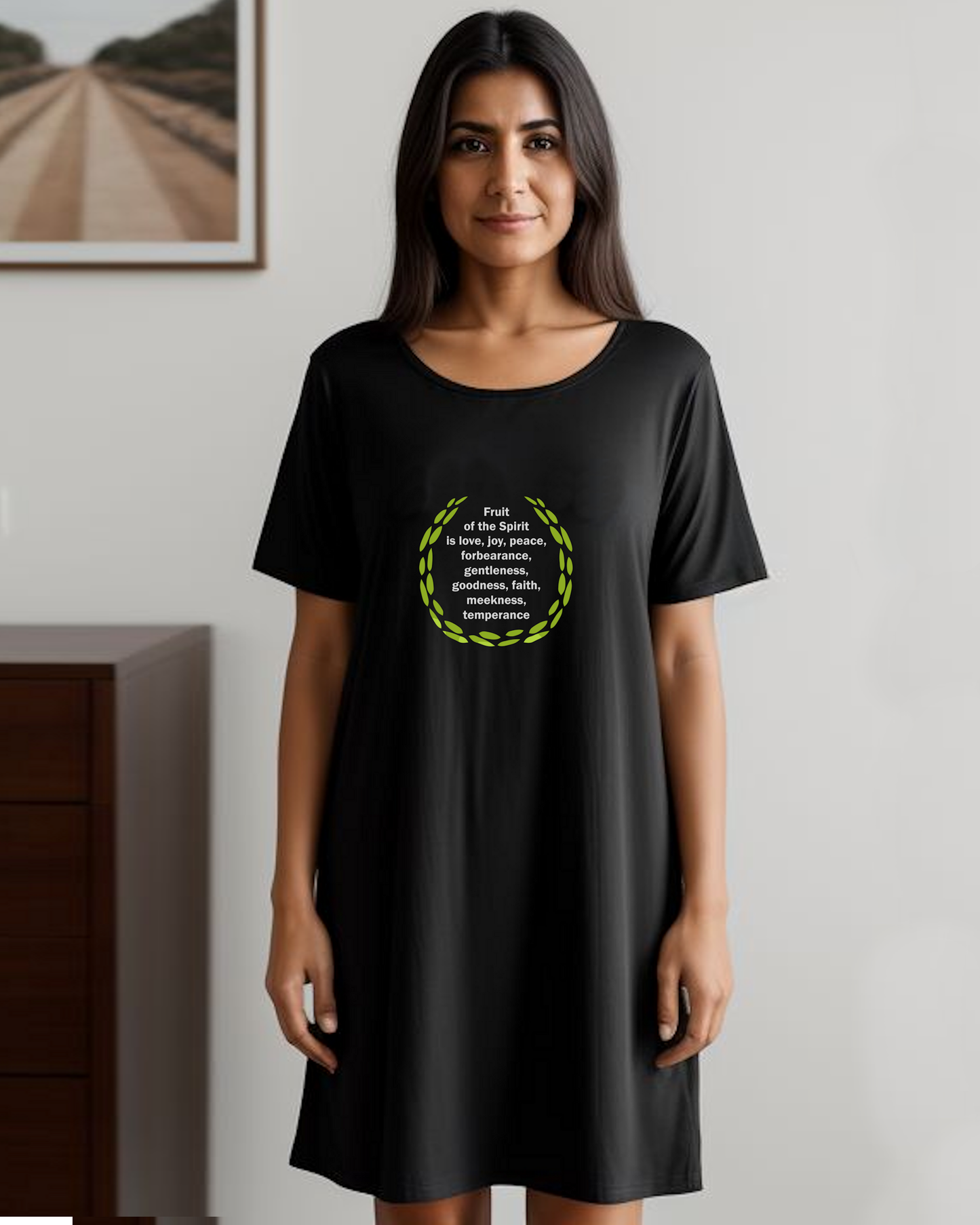 A woman in a bedroom wearing a black t-shirt dress with a logo stating the fruit of the Holy Spirit in white text surrounded by green leaves.