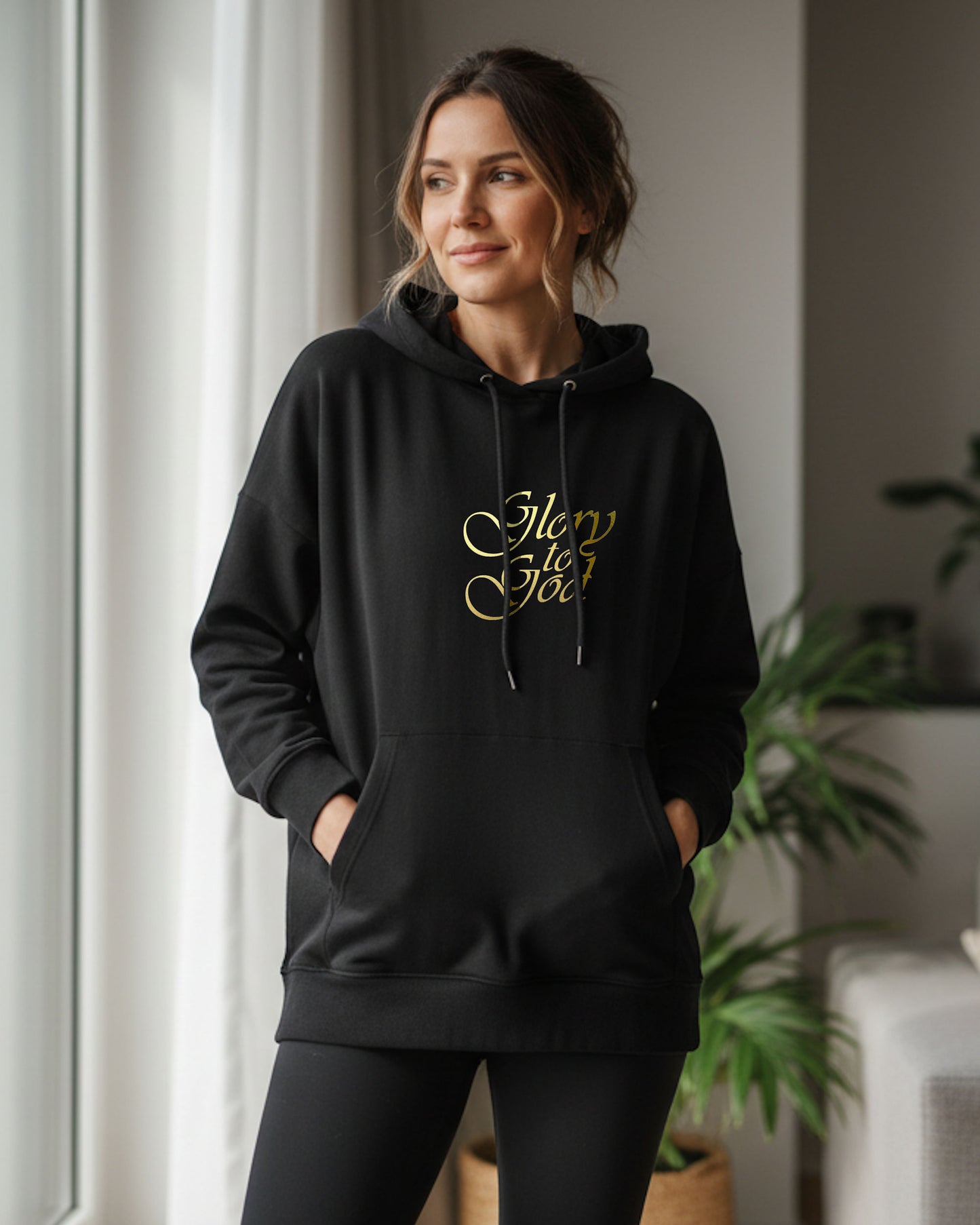 A woman standing indoors wearing a black hoodie with a gold Heavenly Dreamwear "Glory to God" text logo printed on front. 