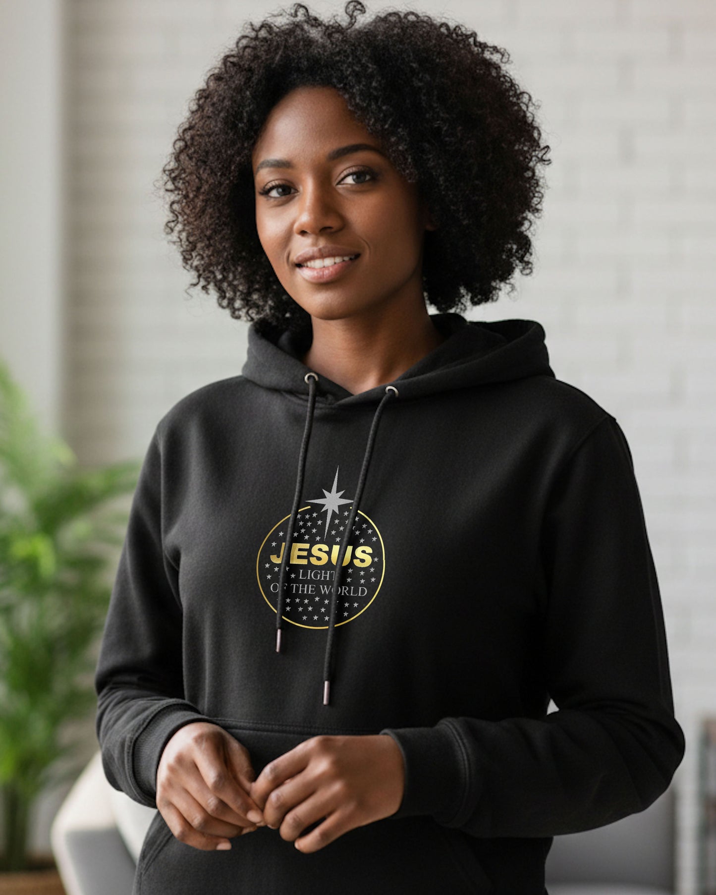 Woman wearing a black hoodie with a silver and gold graphic and text design, standing indoors against a neutral background.