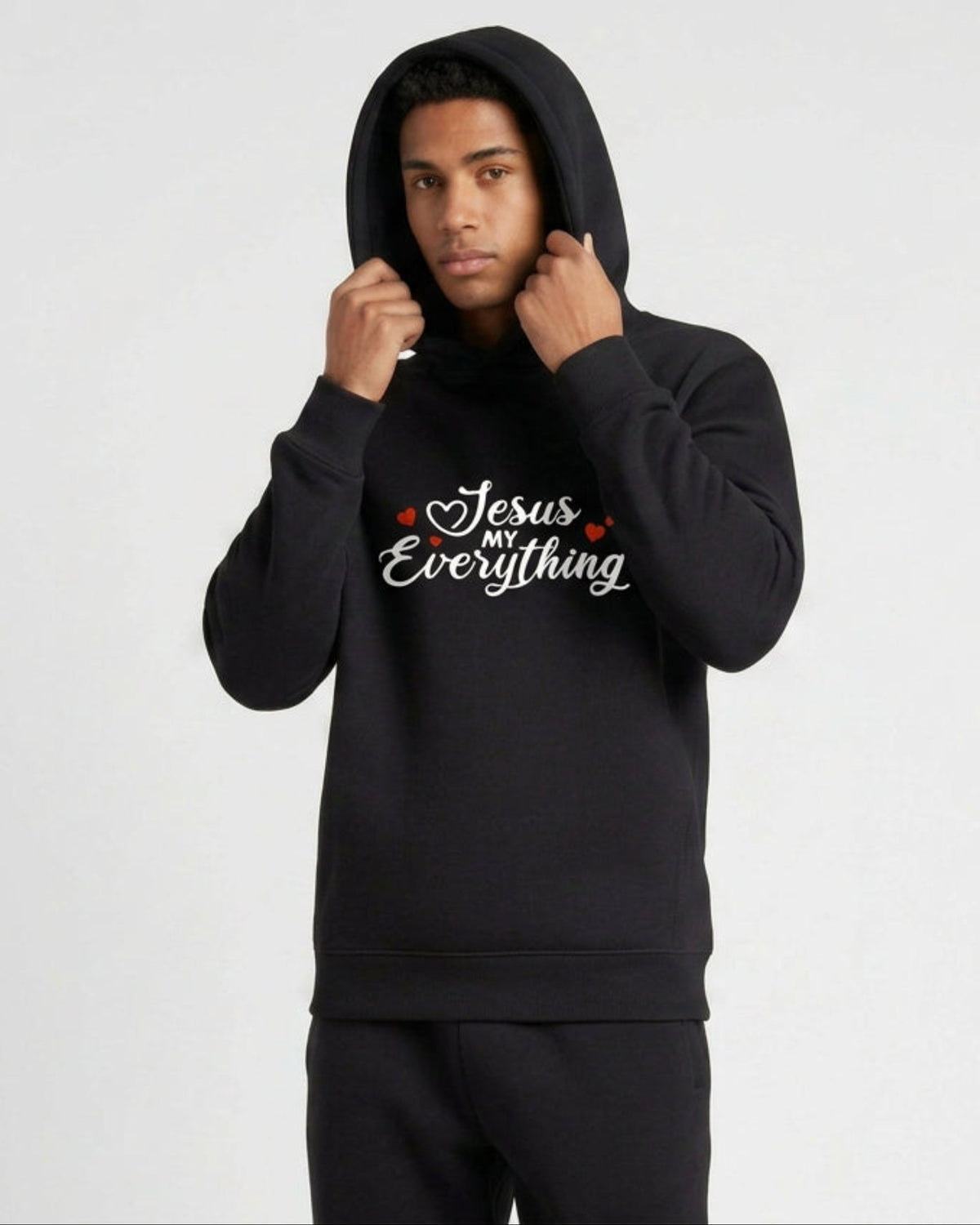 A man wearing a black hoodie with a Christian faith inspired text logo saying "Jesus My Everyhing" designed by Heavenly Dreamwear printed on the front against a white background
