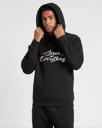 A man wearing a black hoodie with a Christian faith inspired text logo saying "Jesus My Everyhing" designed by Heavenly Dreamwear printed on the front against a white background