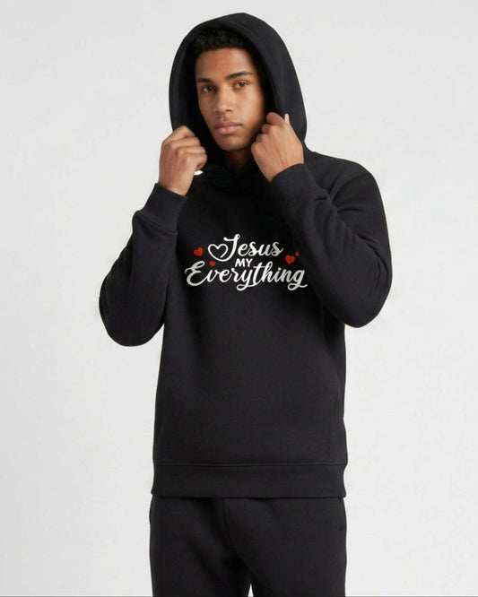 A man wearing a black hoodie with a Christian faith inspired text logo saying "Jesus My Everyhing" designed by Heavenly Dreamwear printed on the front against a white background