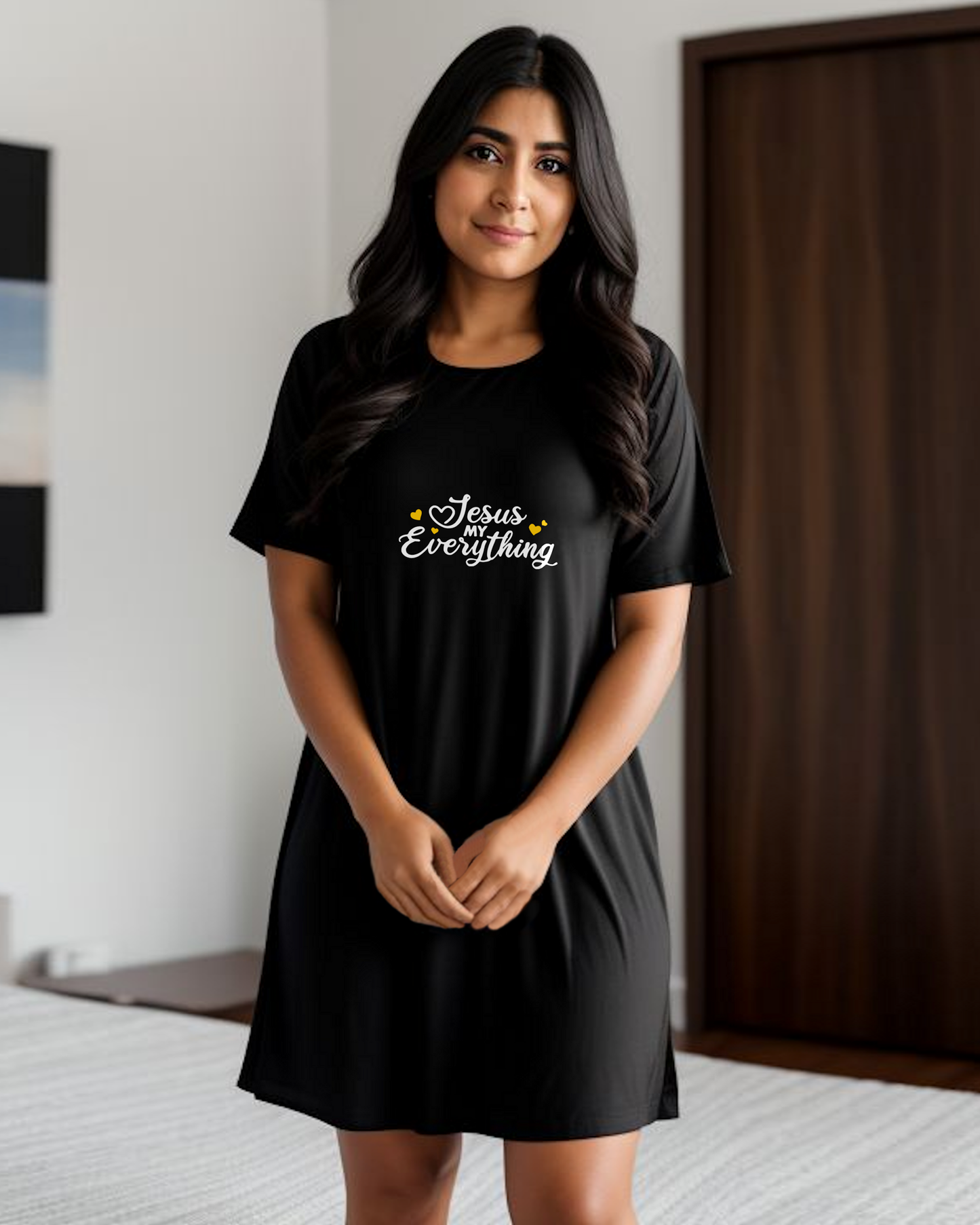 A woman in a bedroom wearing a black Heavenly Dreamwear dress with a logo made up of white text, "Jesus MY Everything", with yellow hearts beside it, printed on the front.