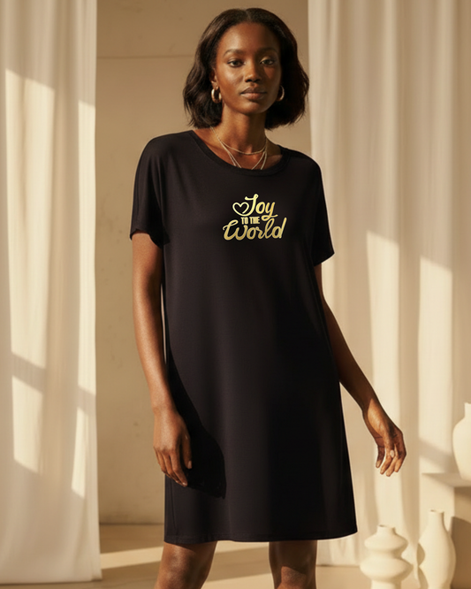 A woman modeling a black short-sleeve shift dress with 'Joy to the World' printed in gold script on the chest, set against a warm, sunlit background with white drapes.