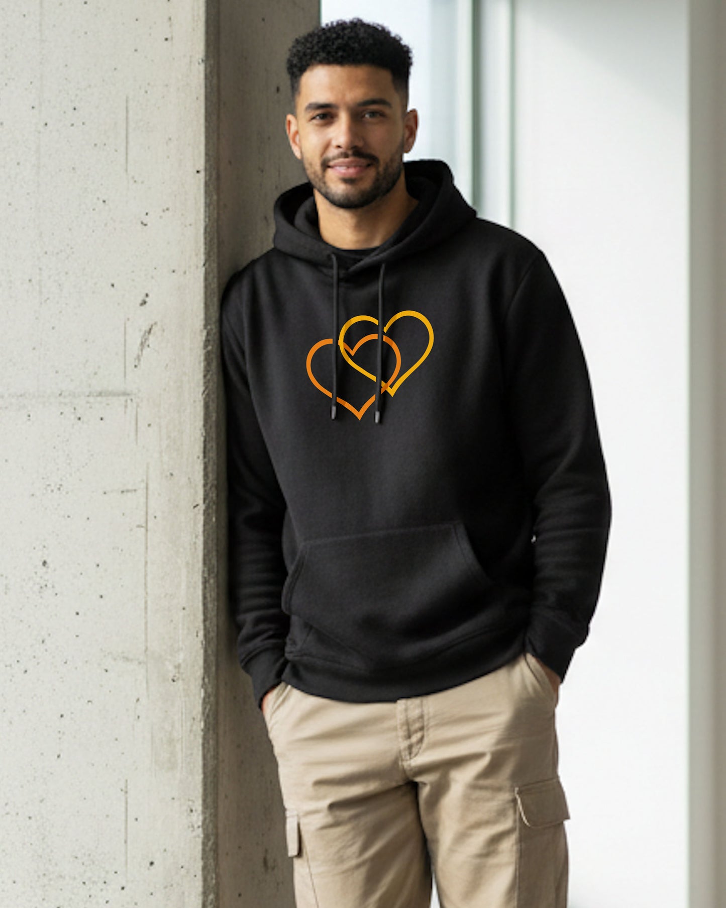 Man wearing a black hoodie with an orange and yellow heart design, leaning against a light-colored wall.