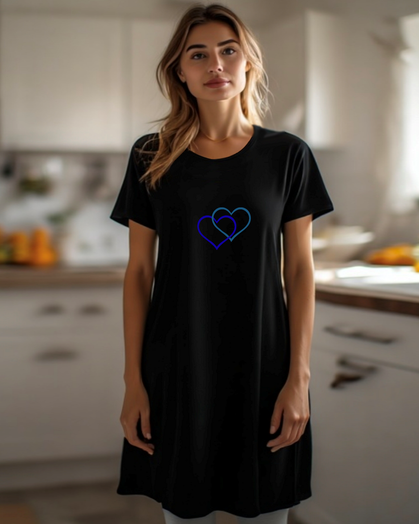 A woman stands in a kitchen, dressed in a black t-shirt dress featuring the Heavenly Dreamwear logo of two intertwined heart shapes printed on the front.