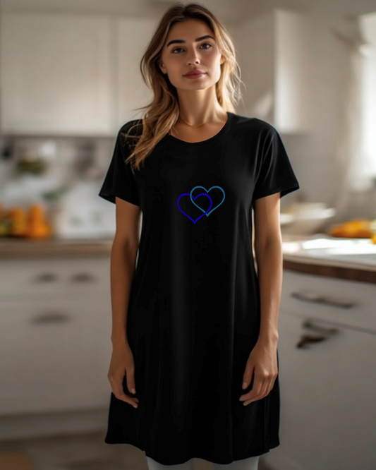 A woman stands in a kitchen, dressed in a black t-shirt dress featuring the Heavenly Dreamwear logo of two intertwined heart shapes printed on the front.