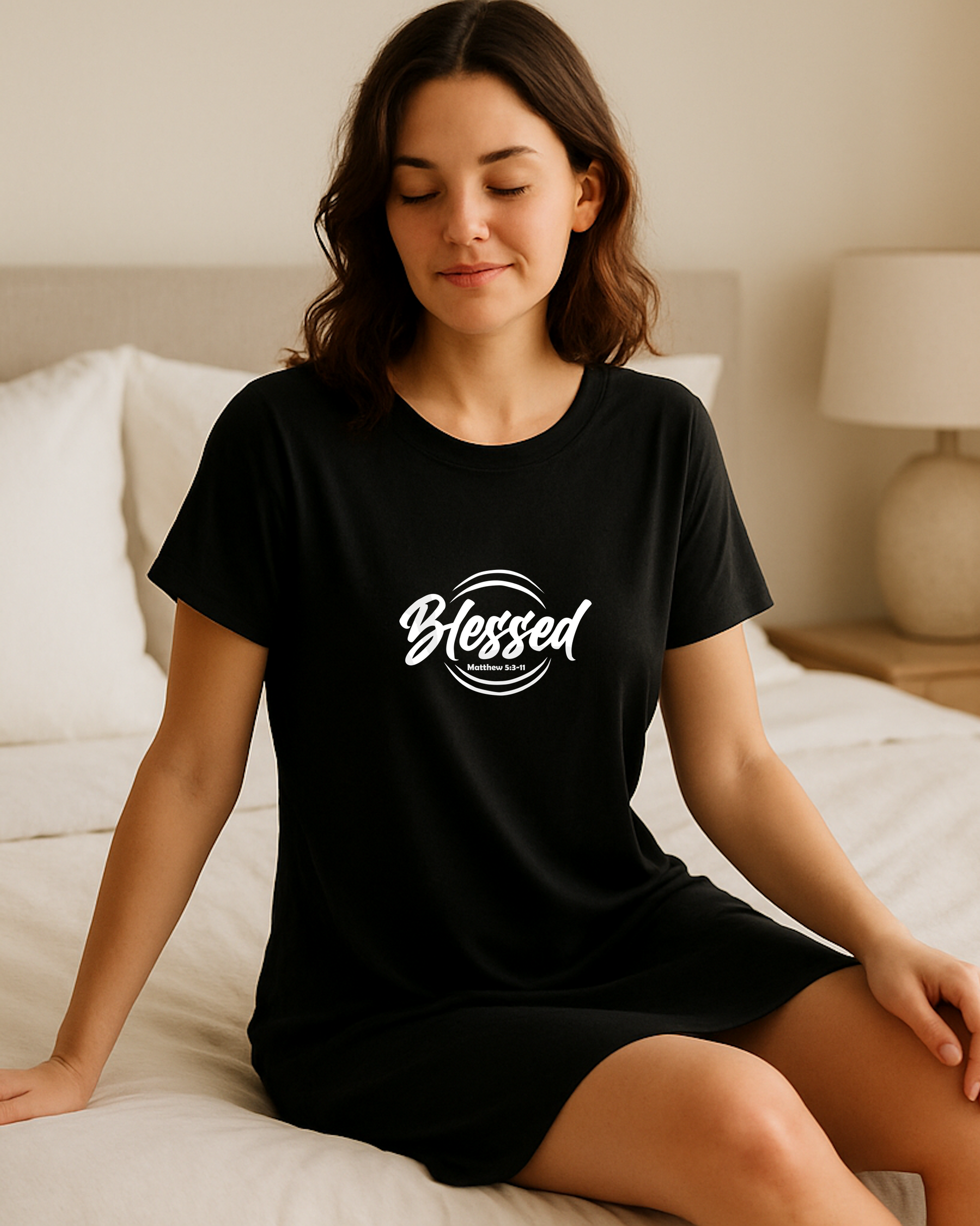 A woman on a bed wearing a black Heavenly Dreamwear t-shirt dress with a white logo comprised of the text "Blessed" on top of a broken double circle with a biblical scripture reference beneath the text.
Photo model: Shopify