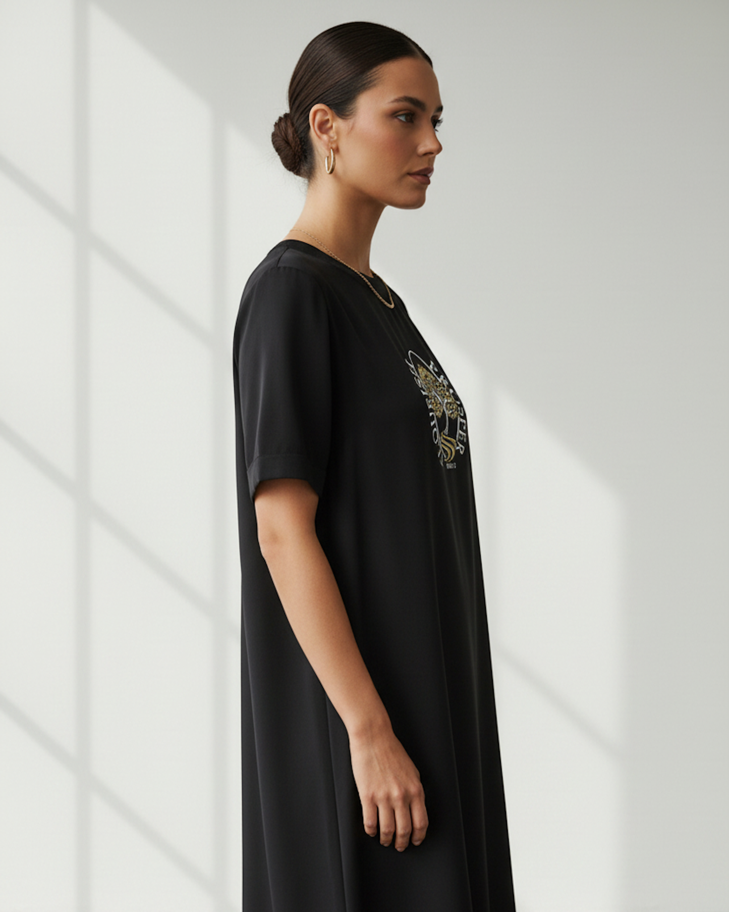 Side view of a woman wearing a black dress with gold and silver logo in a bright room.