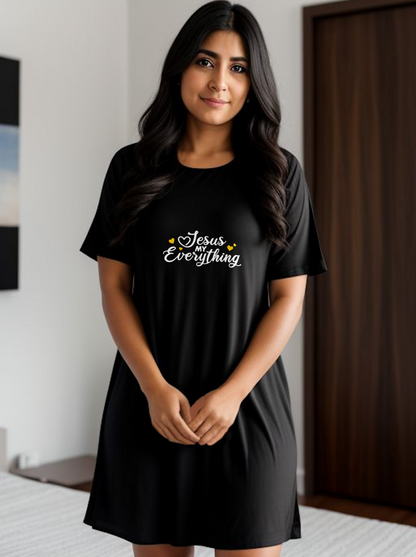 A woman wearing a black short-sleeve t-shirt dress with 'Jesus Everything' printed in white with orange hearts on the chest, standing in a modern room with a dark wood door in the background.