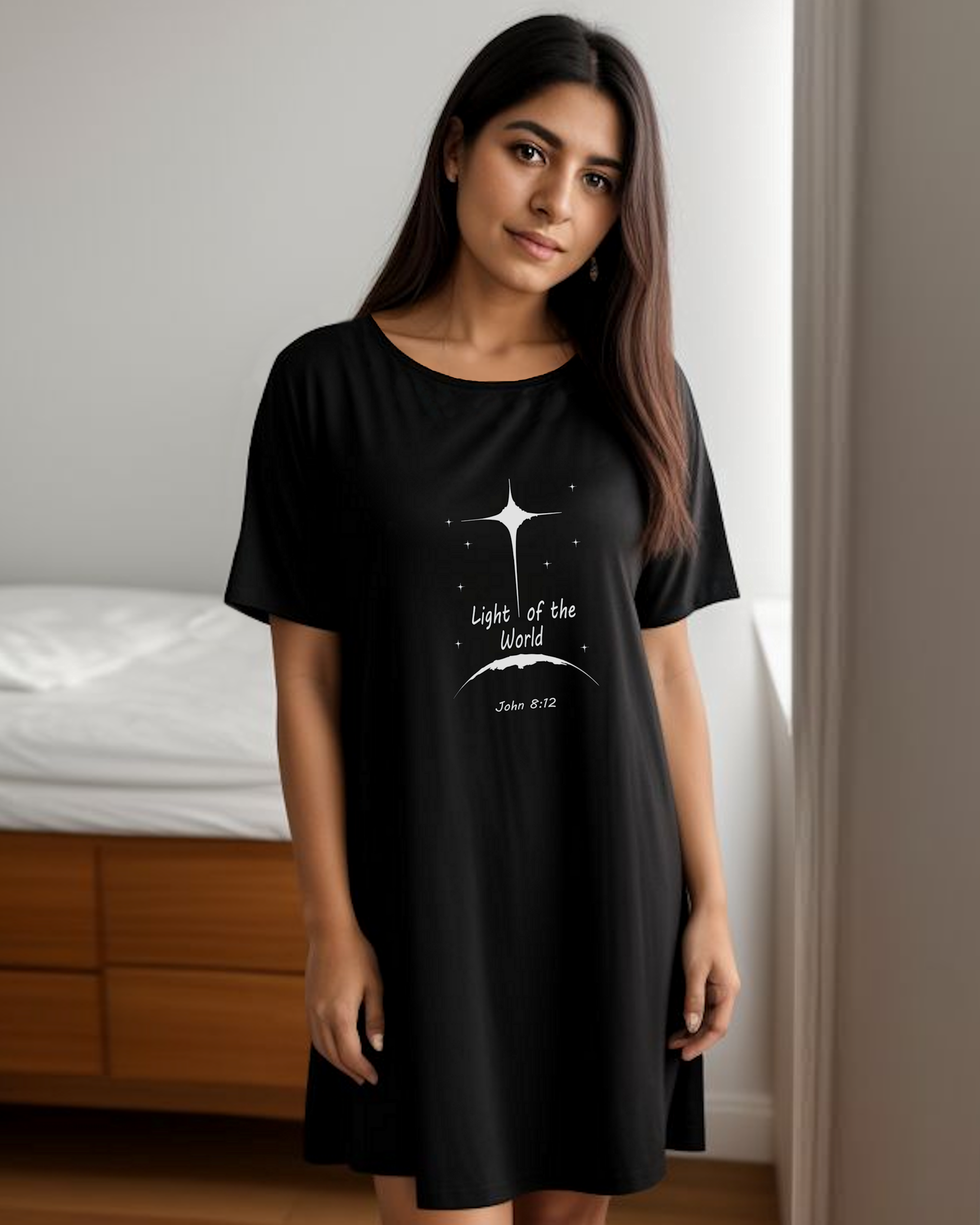 A woman standing in a bedroom wearing a black t-shirt dress with a white Heavenly Dreamwear logo that says "Light of the World" printed on the front.