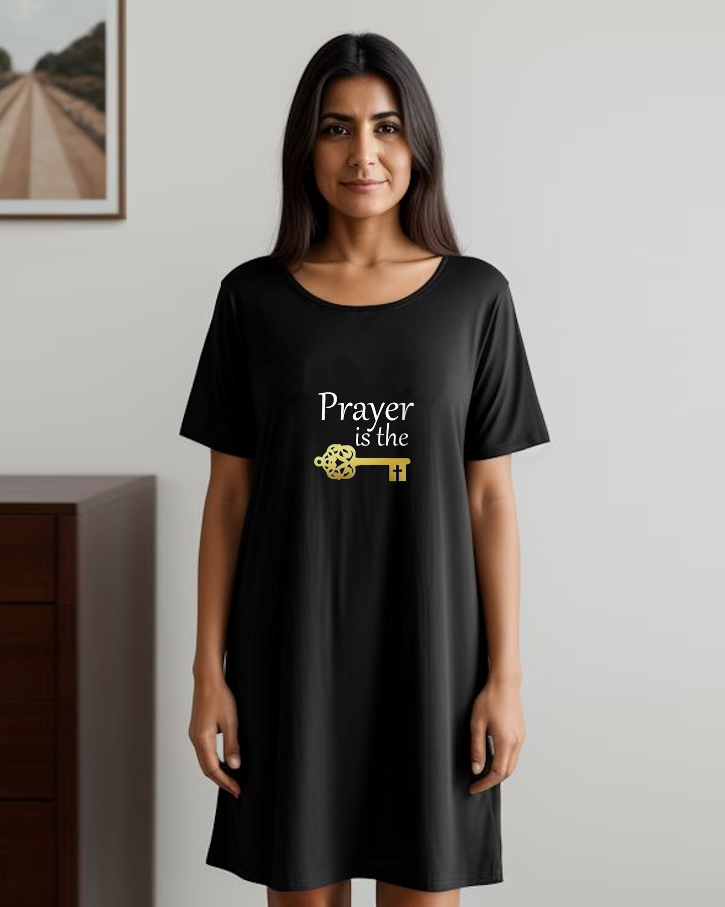 A woman is wearing a black cotton t-shirt dress featuring a design by Heavenly Dreamwear.  The design includes white text that reads, "Prayer is the," followed by a gold key symbol.