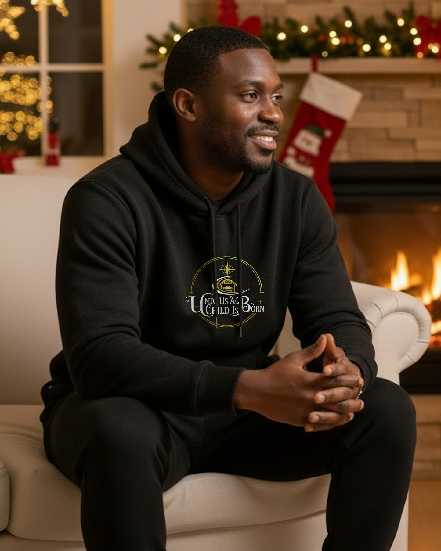 A man wearing a black hoodie that has the white text, "Unto Us A Child Is Born" within a gold logo, in Christmas setting room with a fireplace.