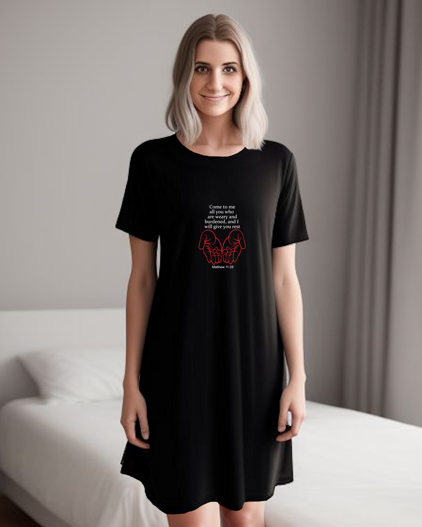 A woman in a bedroom wearing a black Heavenly Dreamwear t-shirt dress with a white "come to me all you who are weary" text with two red hands facing palm up logo printed on the front.