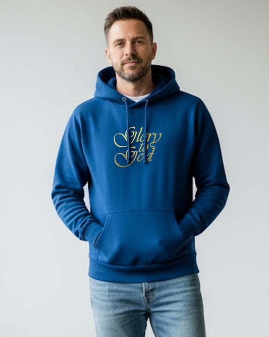 Man standing indoors wearing a blue hoodie with a gold Heavenly Dreamwear "Glory to God" text logo printed on front. 