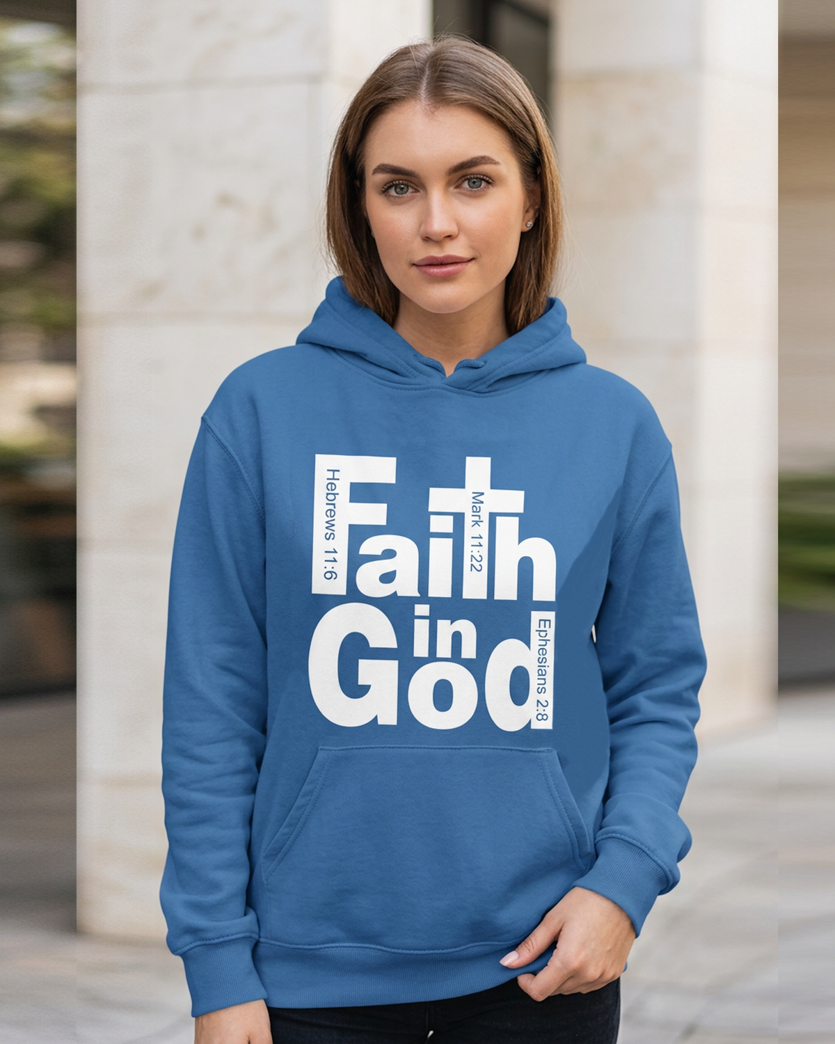 A woman wearing a medium blue hoodie with a white graphic text print reading 'Faith in God' and Bible verse references, standing outdoors in front of a light-colored stone column.