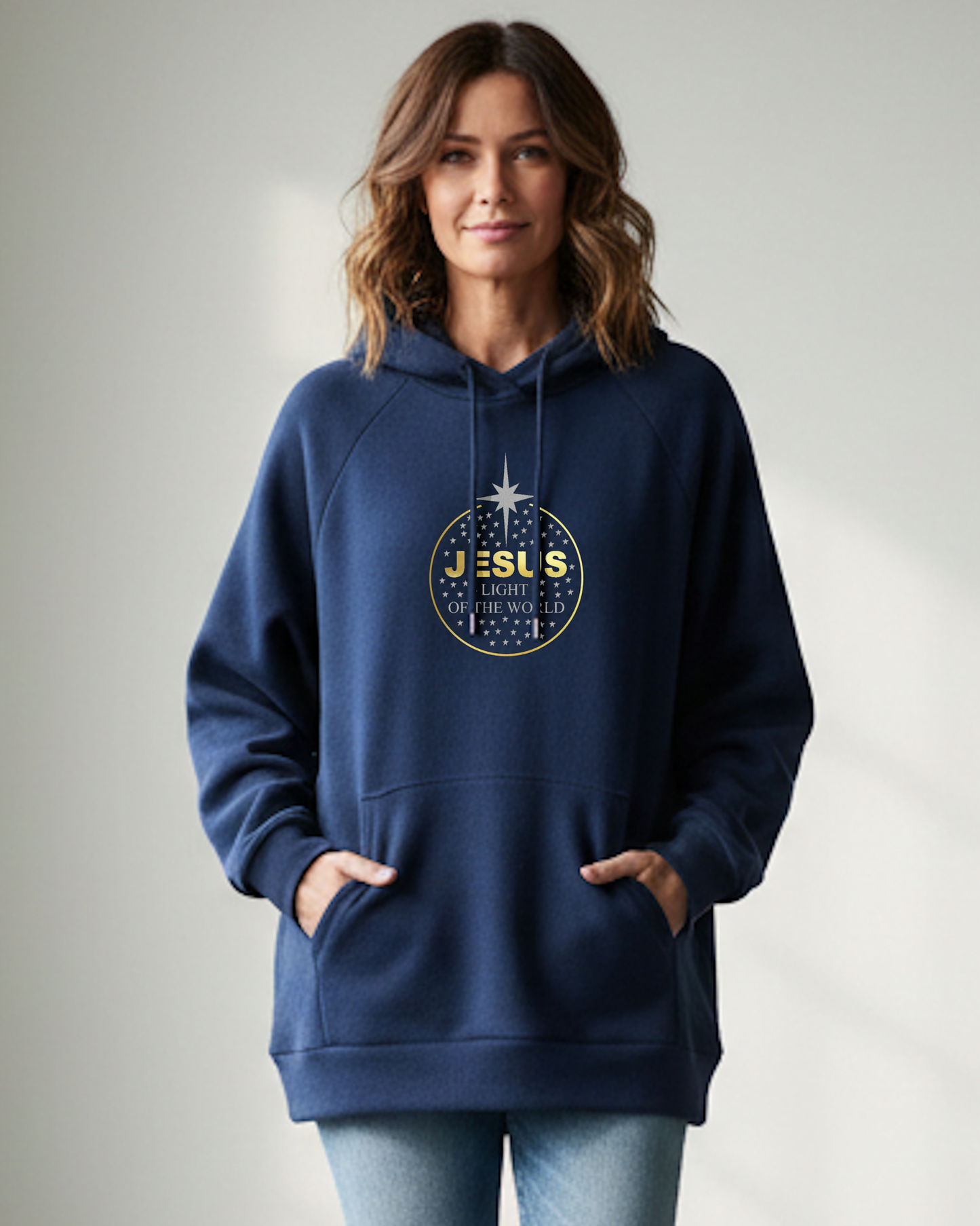 A woman standing indoors wearing a blue hoodie with a gold/silver text and graphic logo. The text reads 'Jesus light of the World'