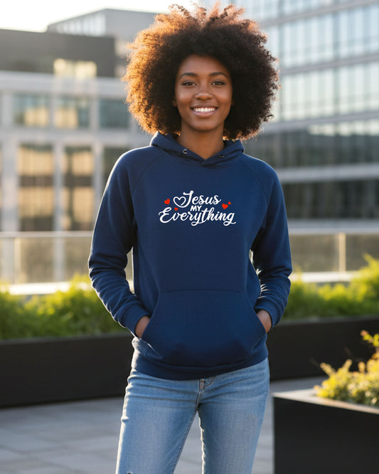 A woman outdoors wearing a blue hoodie with a Christian faith inspired text logo saying "Jesus My Everyhing" designed by Heavenly Dreamwear printed on the front.