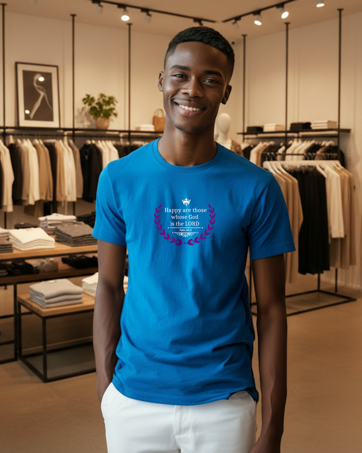 A smiling man wearing a bright blue graphic t-shirt and white pants stands inside a clothing store. The t-shirt features a purple and white graphic with a maple leaf and the text 'Happy are those whose God is the LORD'.