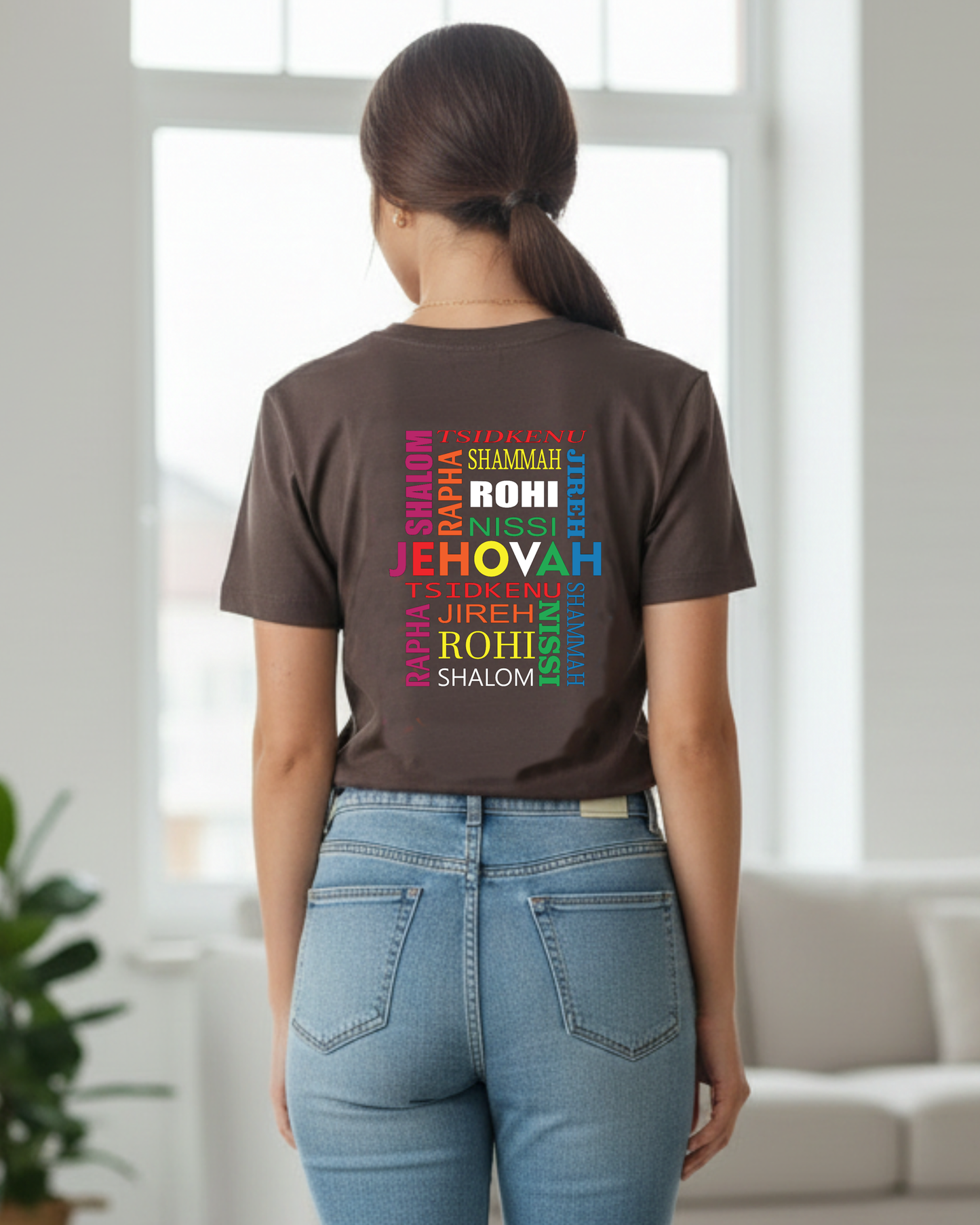 A woman wearing a dark brown t-shirt with a colorful rainbow text graphic on the back that reads 'JEHOVAH, TSIDKENU, SHAMMAH, SHALOM, JIREH, RAPHA NISSI, ROHI' paired with light blue jeans, standing in a bright, modern room.