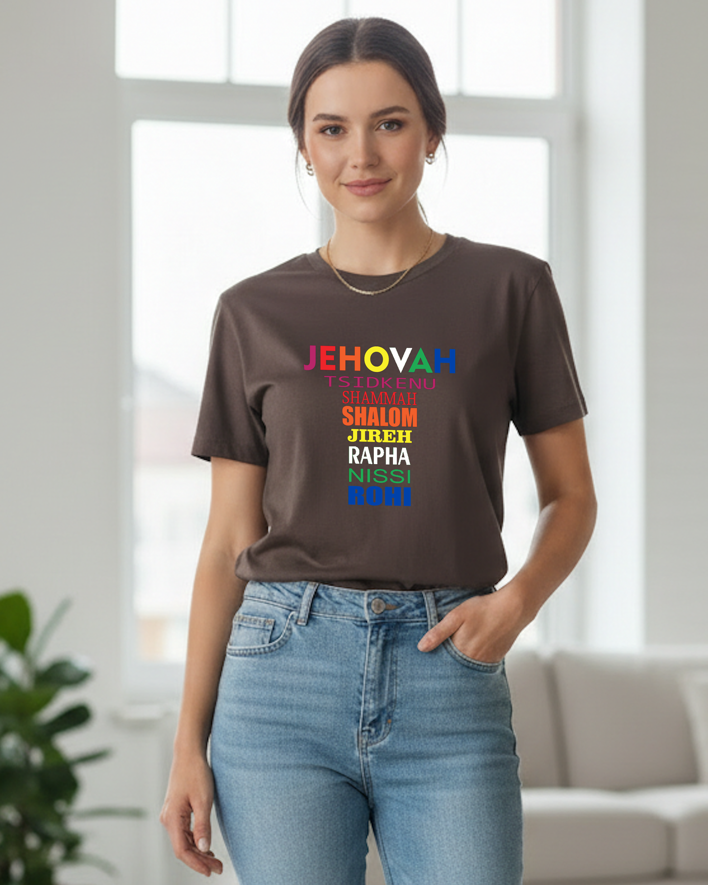 A woman wearing a dark brown t-shirt with a colorful rainbow text graphic that reads 'JEHOVAH, TSIDKENU, SHAMMAH, SHALOM, JIREH, RAPHA NISSI, ROHI' paired with light blue jeans, standing in a bright, modern room.