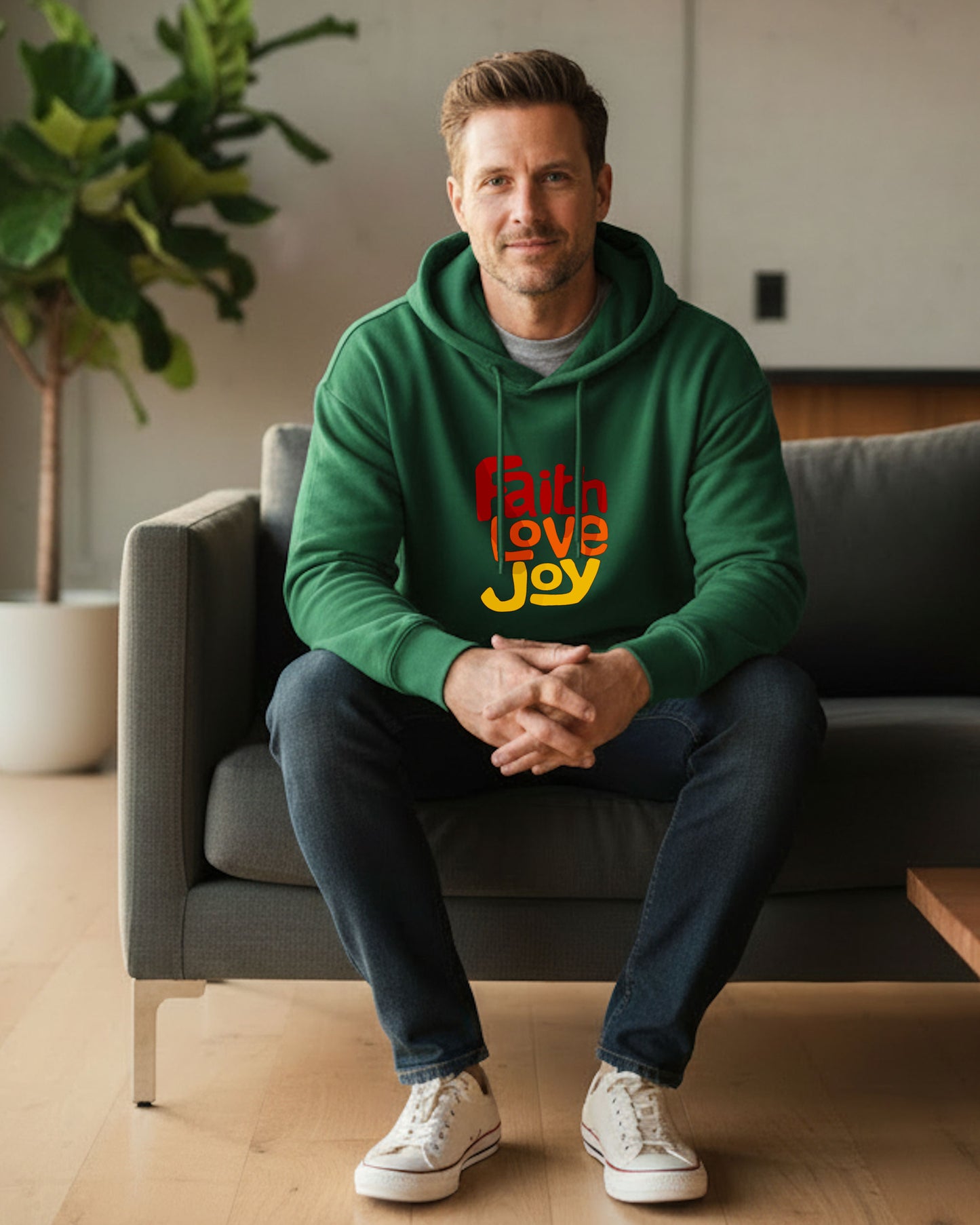 Man wearing a green hoodie with 'Faith Love Joy' text, sitting on a couch in a living room.